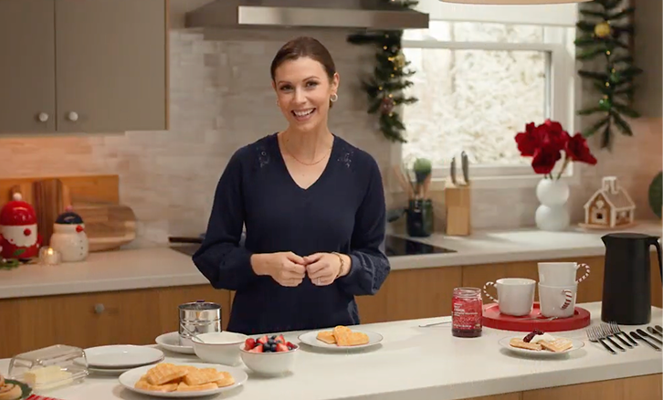 A woman standing in a kitchen decorated for Christmas, smiling at the camera, with plates, cups, and snacks on the counter in front of her.