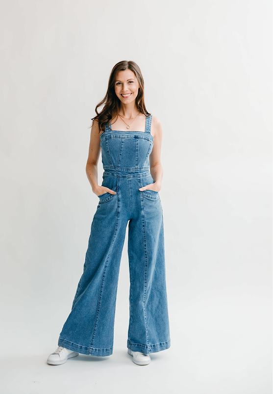 Young woman with brown hair smiling, wearing a denim jumpsuit and white sneakers, standing against a plain white background.