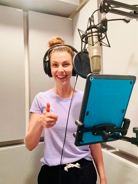 A young woman with a bun hairstyle wearing headphones, standing in front of a microphone setup with a pop filter and a tablet, smiling and giving a thumbs-up.