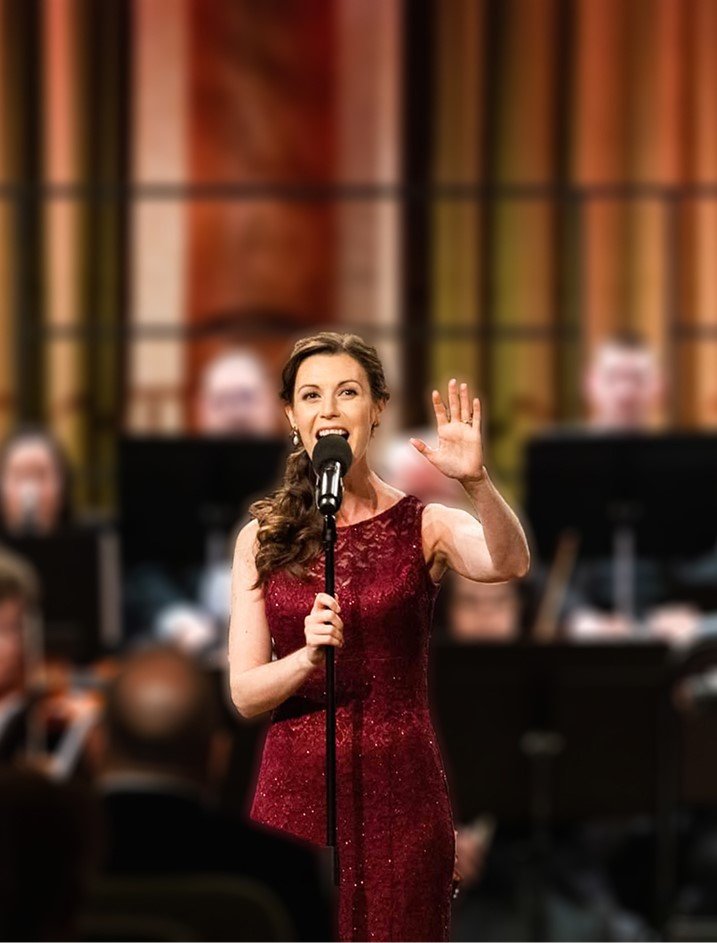 A woman in a red dress speaking or singing into a microphone at an event with a blurred orchestra or group of musicians in the background.