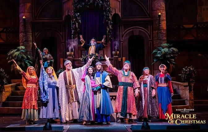Cast performing on stage during a Christmas-themed theater production, with actors dressed in colorful, medieval-style costumes, in front of a castle-like set with a figure dressed as a king or prophet in the background.