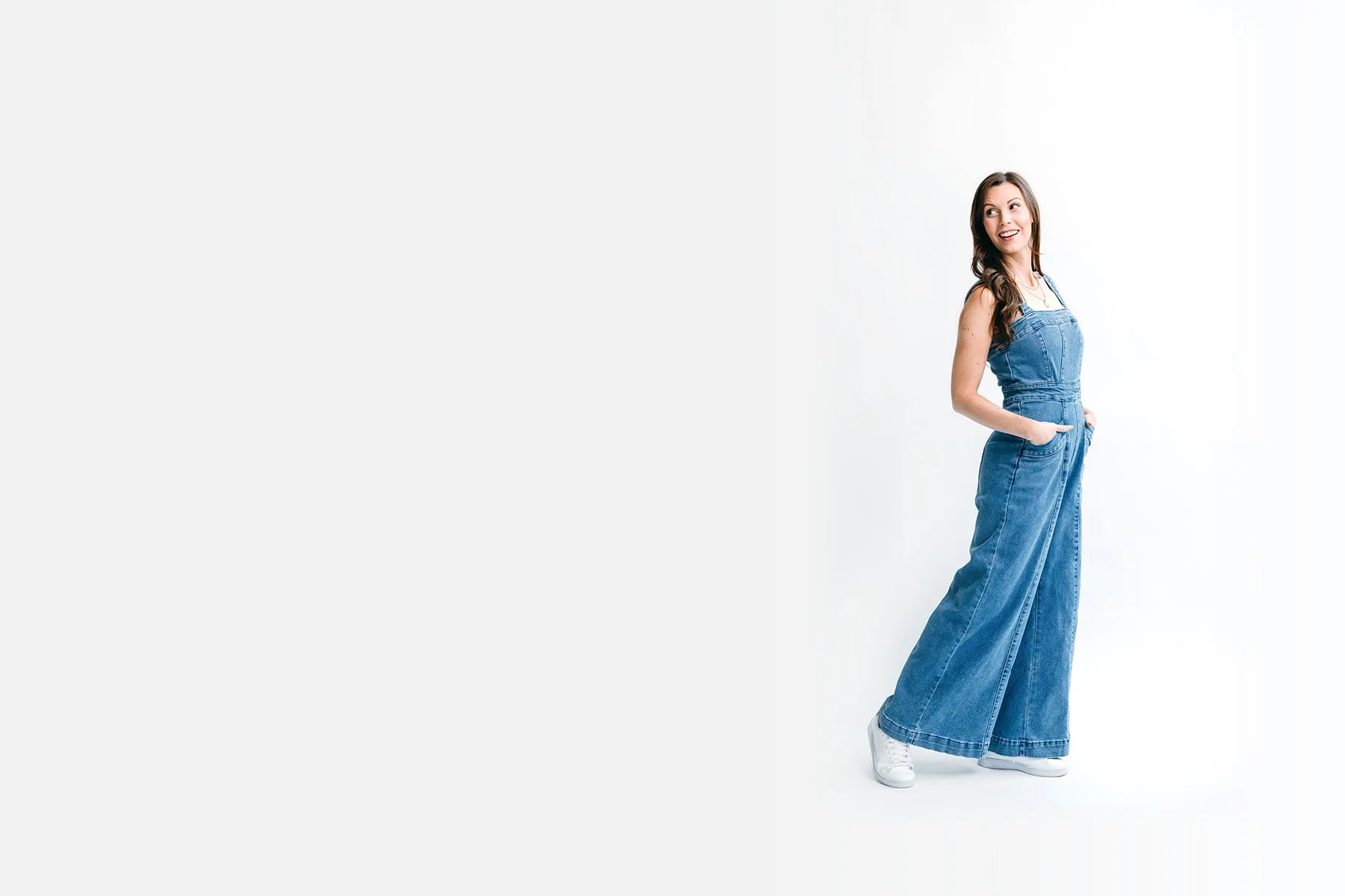 A woman with long brown hair wearing a denim jumpsuit and white sneakers, smiling and looking to her right, standing against a plain white background.