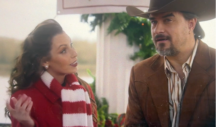 A woman with wavy hair, wearing a red sweater and a red-and-white striped scarf, looks at a man with a beard and mustache, wearing a brown jacket and a cowboy hat. They are outdoors with a blurred background of greenery and a white column.