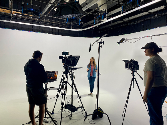 A video production or photography studio with a woman posing in front of a white backdrop, two crew members working with cameras and equipment, and lighting and mic setups overhead.