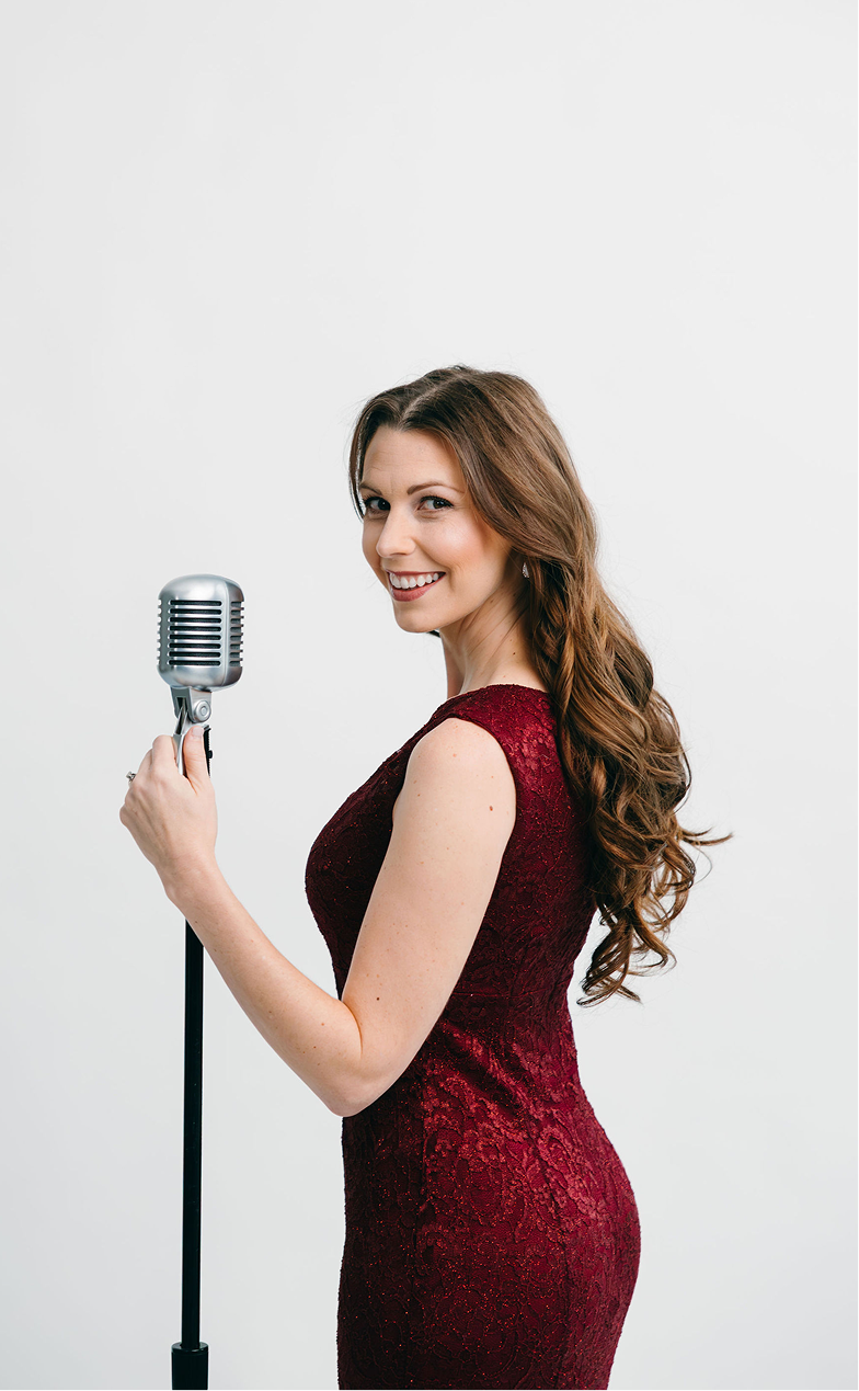 A woman with long, curly brown hair wearing a red lace dress, holding a vintage microphone and smiling at the camera.