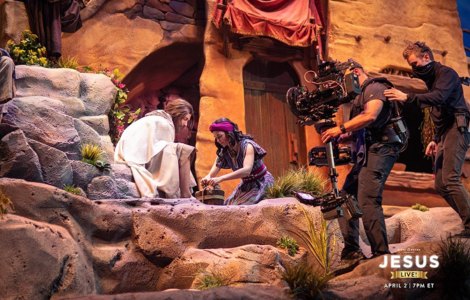 Actors filming a scene of Jesus with a woman and a girl on a rocky set with desert plants, led by a camera operator.