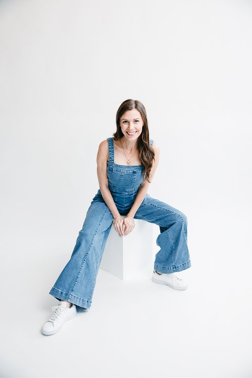 Young woman sitting on a white block against a plain white background, wearing denim overalls and white sneakers, smiling at the camera.