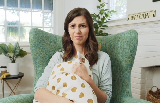 A woman sitting in a green armchair holding a pillow with gold polka dots, looking unsure or worried, in a bright living room with large windows and green plants.