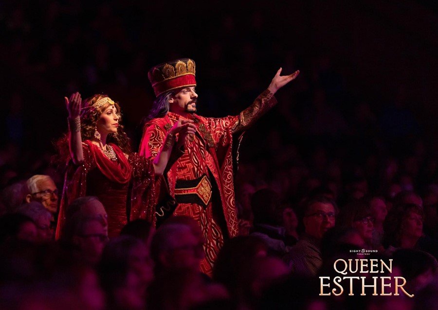 People watching a theatrical performance titled 'Queen Esther,' with actors dressed in elaborate costumes inspired by historical or biblical attire in a darkened theater.