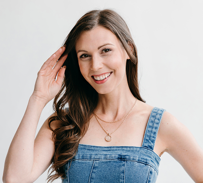 A young woman with long brown hair smiling and touching her hair, wearing a denim sleeveless top and a gold necklace, against a plain light background.