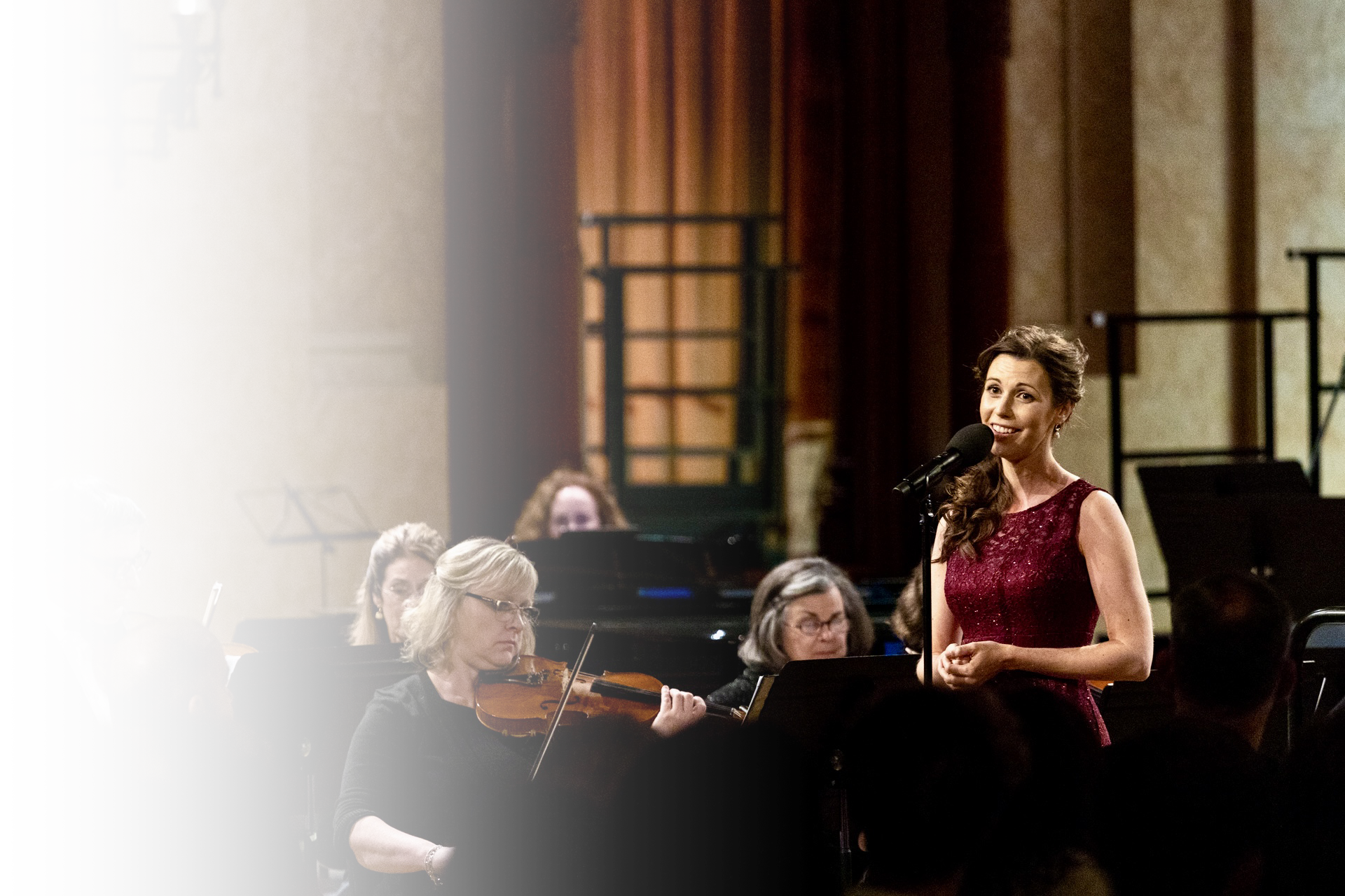 A woman in a red dress performs as a singer with an orchestra accompanying her in a concert hall.