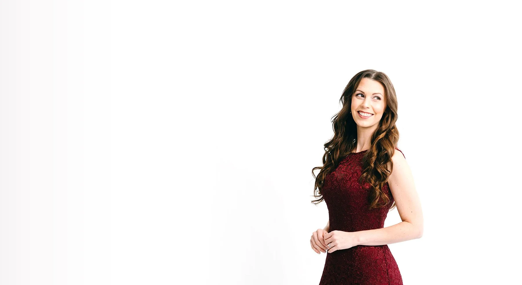 A smiling woman with long, wavy brown hair wearing a sleeveless, burgundy lace dress standing against a plain white background.