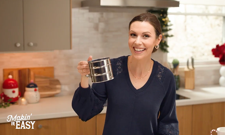 A woman smiling and holding a set of stainless steel measuring cups in a kitchen.