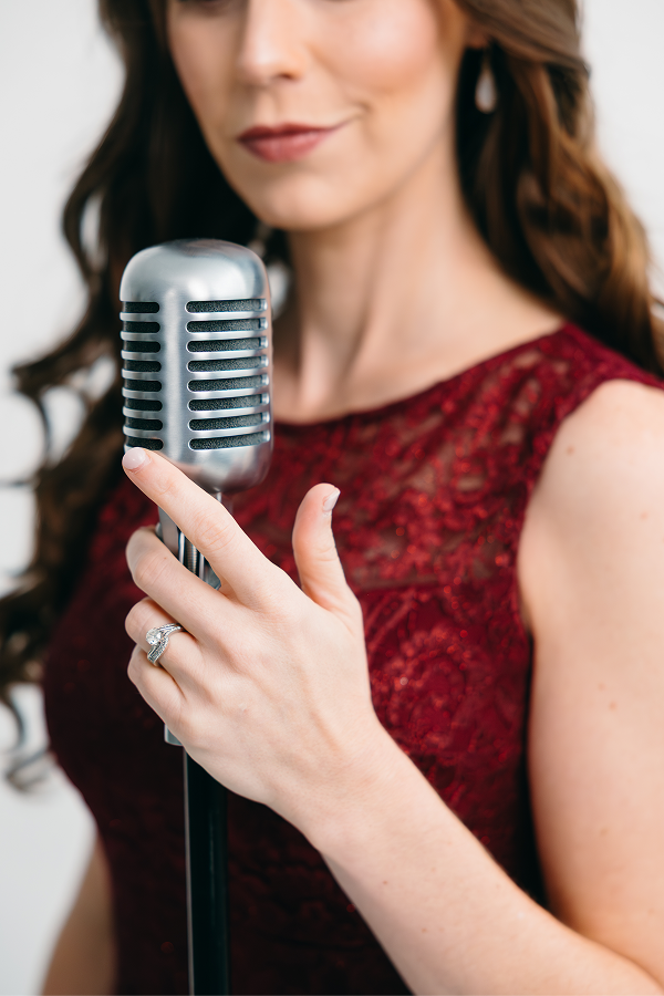 A woman in a red lace dress holding a vintage silver microphone, smiling softly.