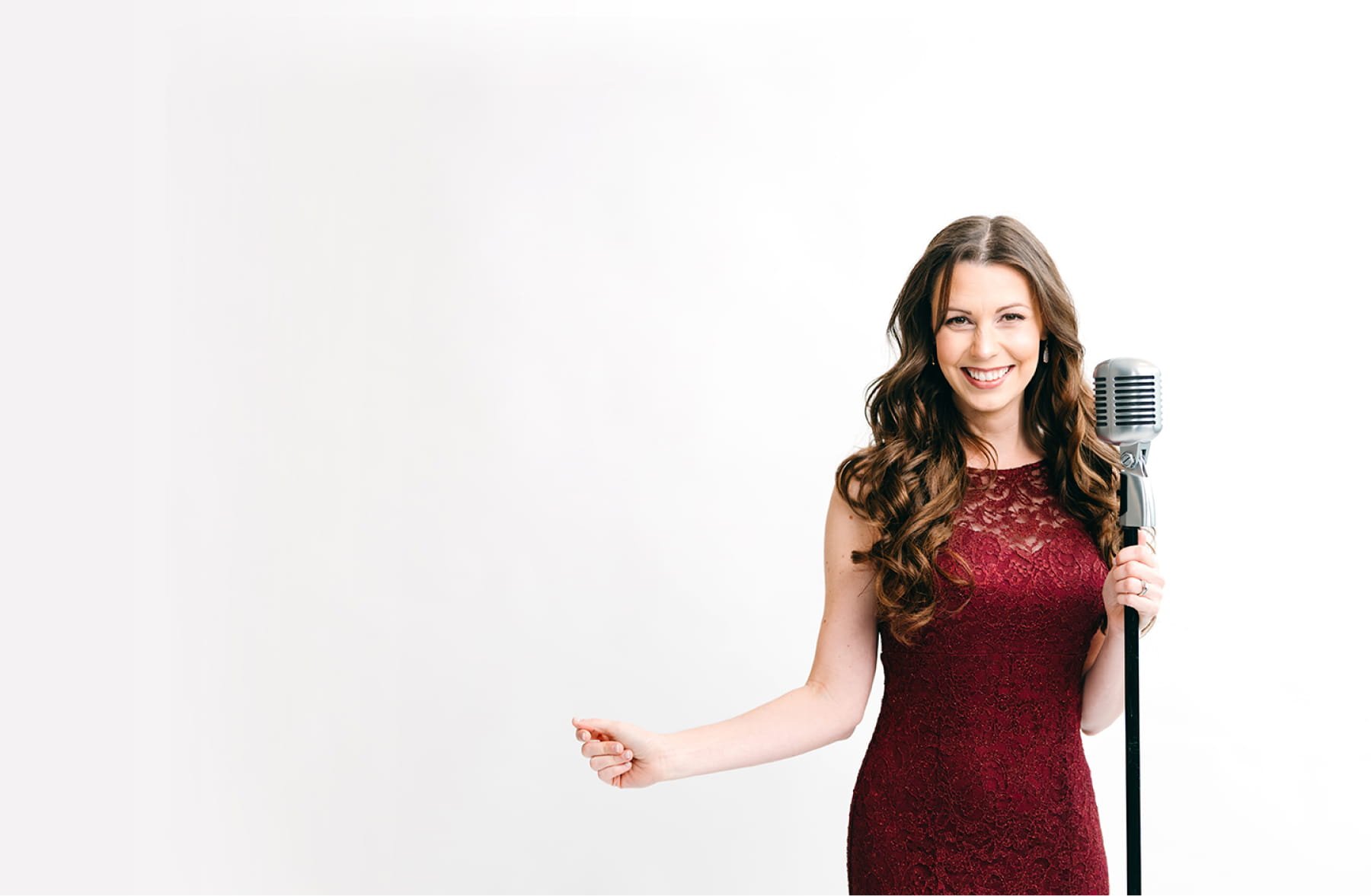 A woman with long curly brown hair wearing a red lace dress, smiling, holding a vintage silver microphone, standing against a plain white background.