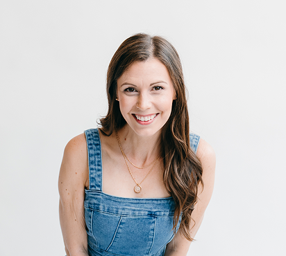 A woman with long brown hair smiling, wearing a denim sleeveless top and a gold necklace, against a white background.