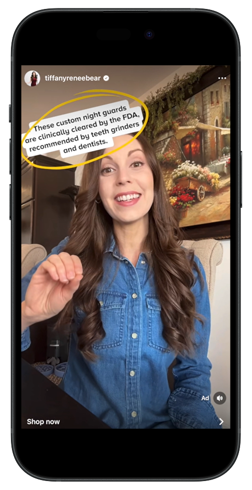 A woman with long brown hair wearing a denim shirt sitting indoors in front of a painting, smiling and gesturing with her right hand. A text overlay indicates she is talking about custom night guards recommended by the FDA, teeth grinders, and dentists.