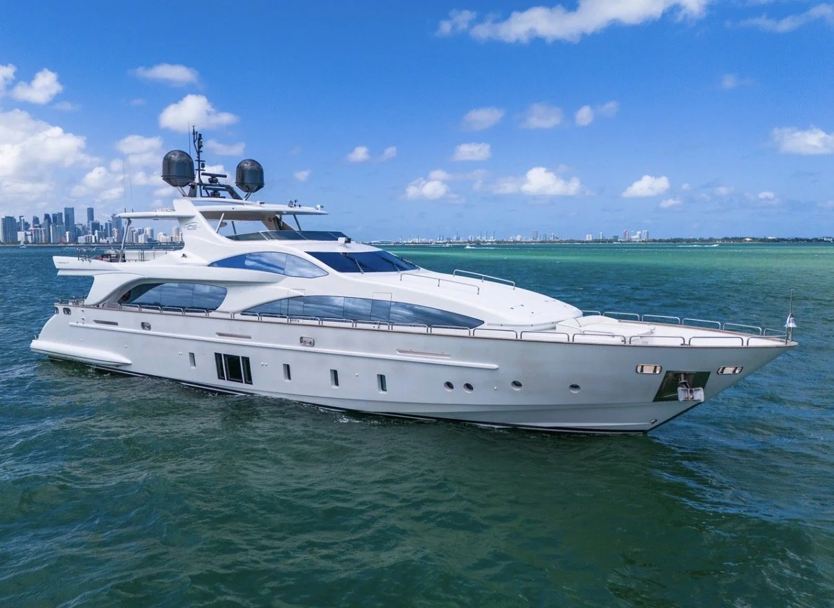 A large white yacht sailing on turquoise water with a city skyline in the background under a partly cloudy sky.