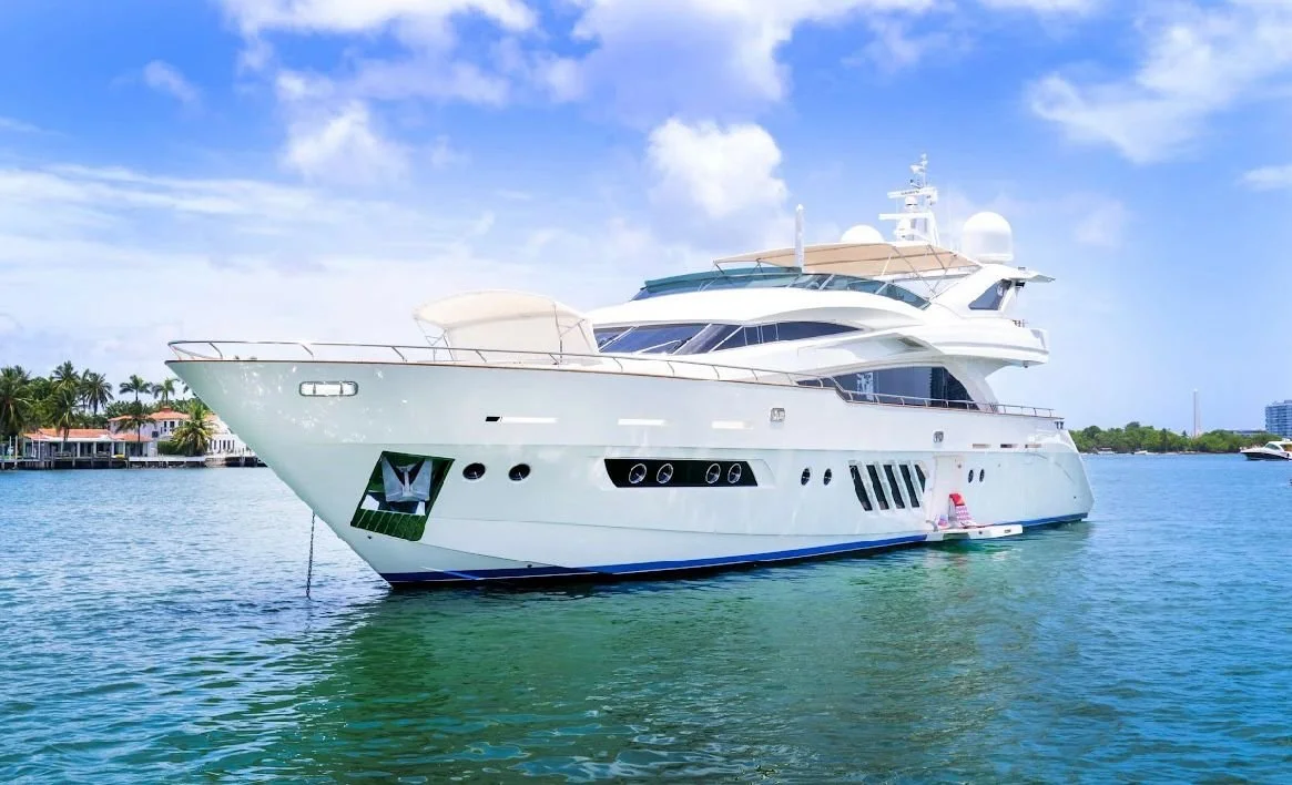 A large white yacht docked in a calm water harbor with palm trees and residential buildings in the background under a partly cloudy sky.