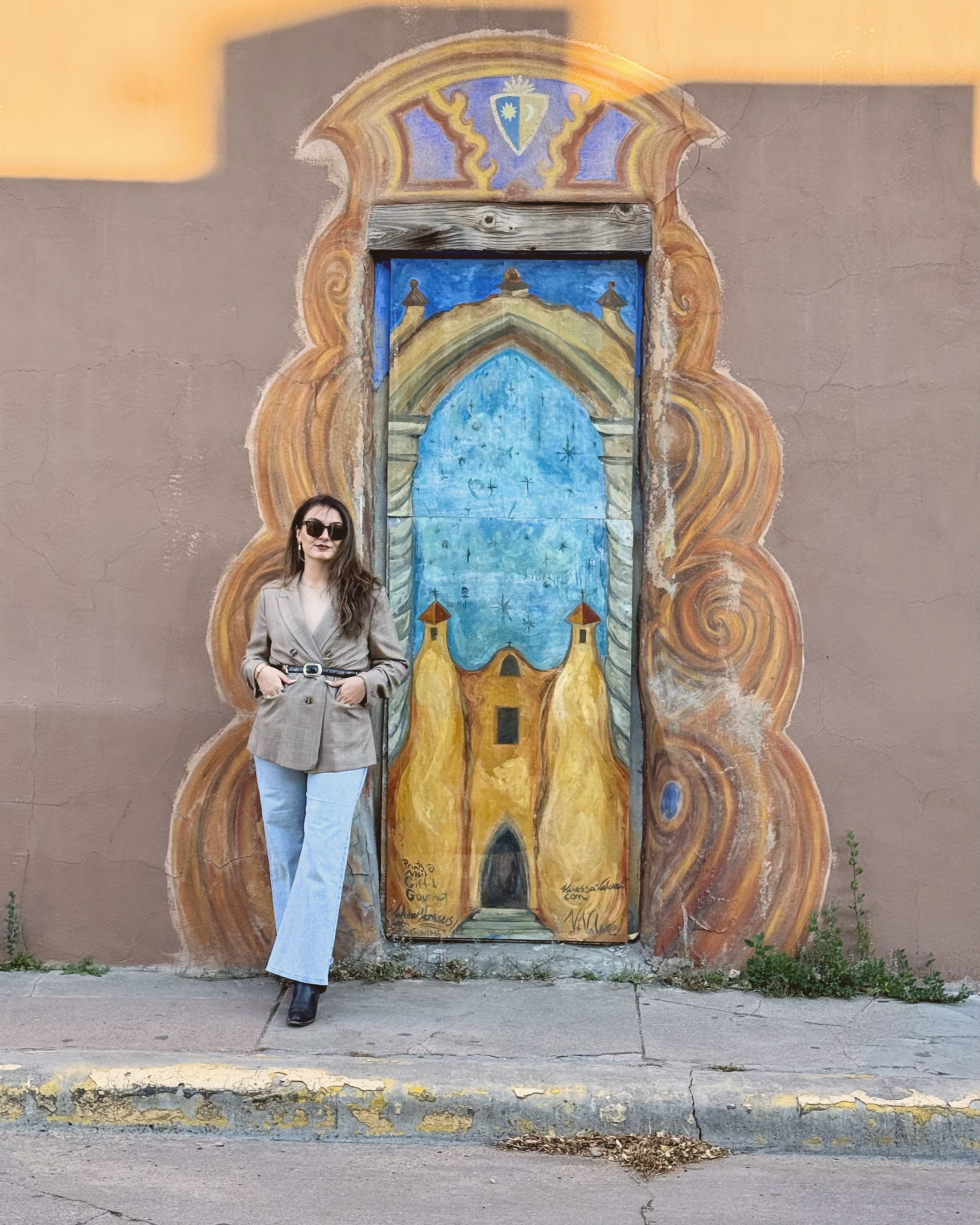 A woman with long hair, glasses, and a beige blazer standing in front of a colorful mural of a door with an ornate frame. The woman is wearing light blue jeans and black boots, with her hands in her pockets.