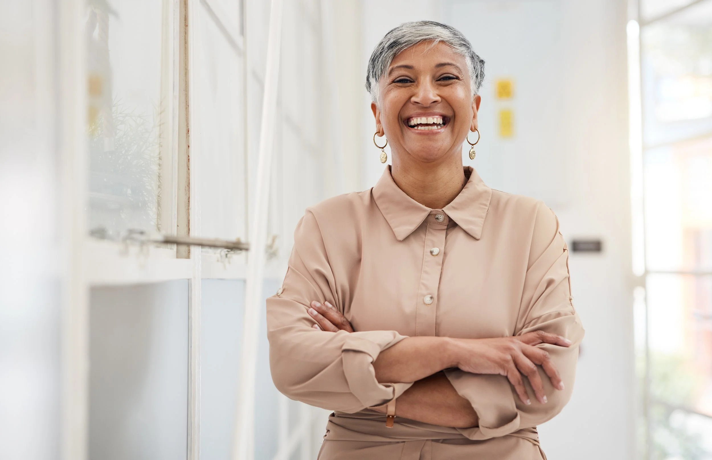 A smiling older woman with short gray hair, wearing a beige shirt with rolled-up sleeves, standing with arms crossed in a bright, modern interior space.