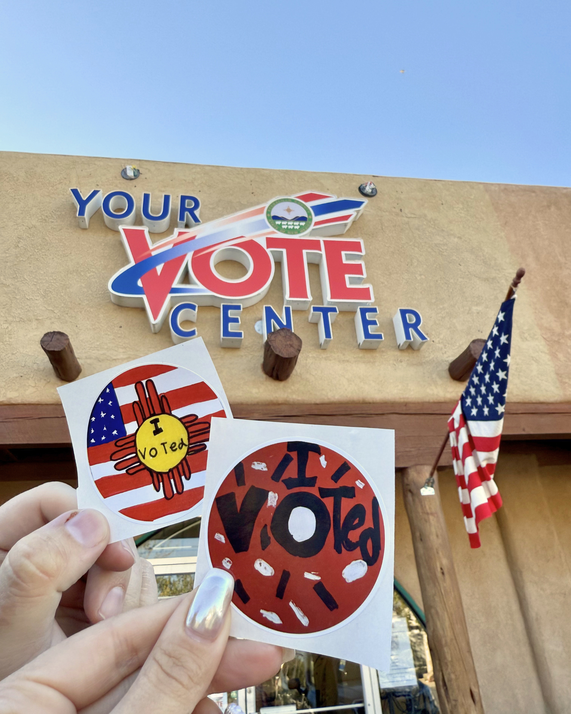 Two hands holding two stickers, one with a peace sign, American flag design, and the words 'I Voted' and another with a red and black circular design and the word 'VOTE.' In the background, a sign reads 'Your Vote Center' with patriotic decorations.