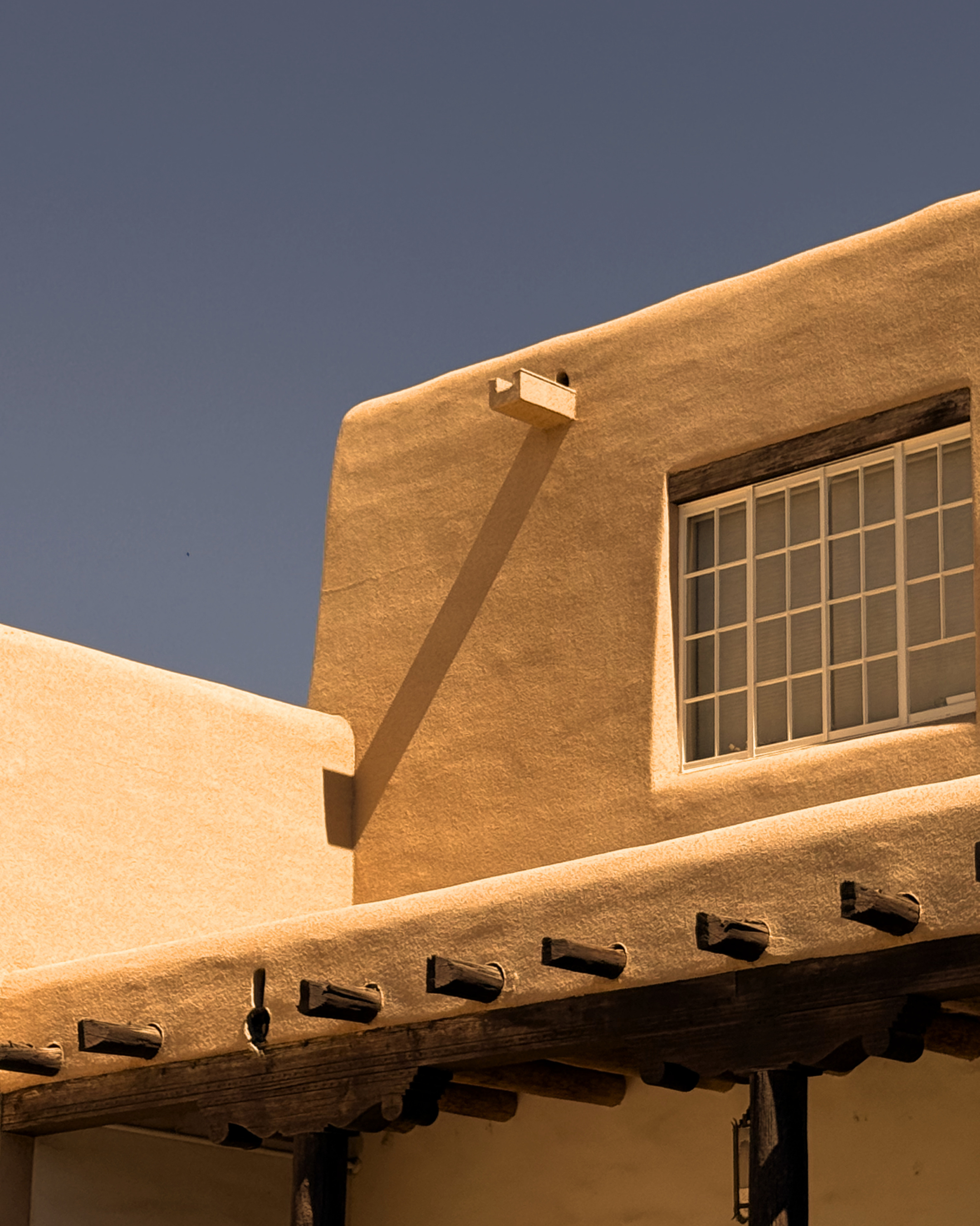 Close-up of a stucco building exterior with a small porch, featuring a grid window and a wooden roof structure with curved edges and decorative brackets.