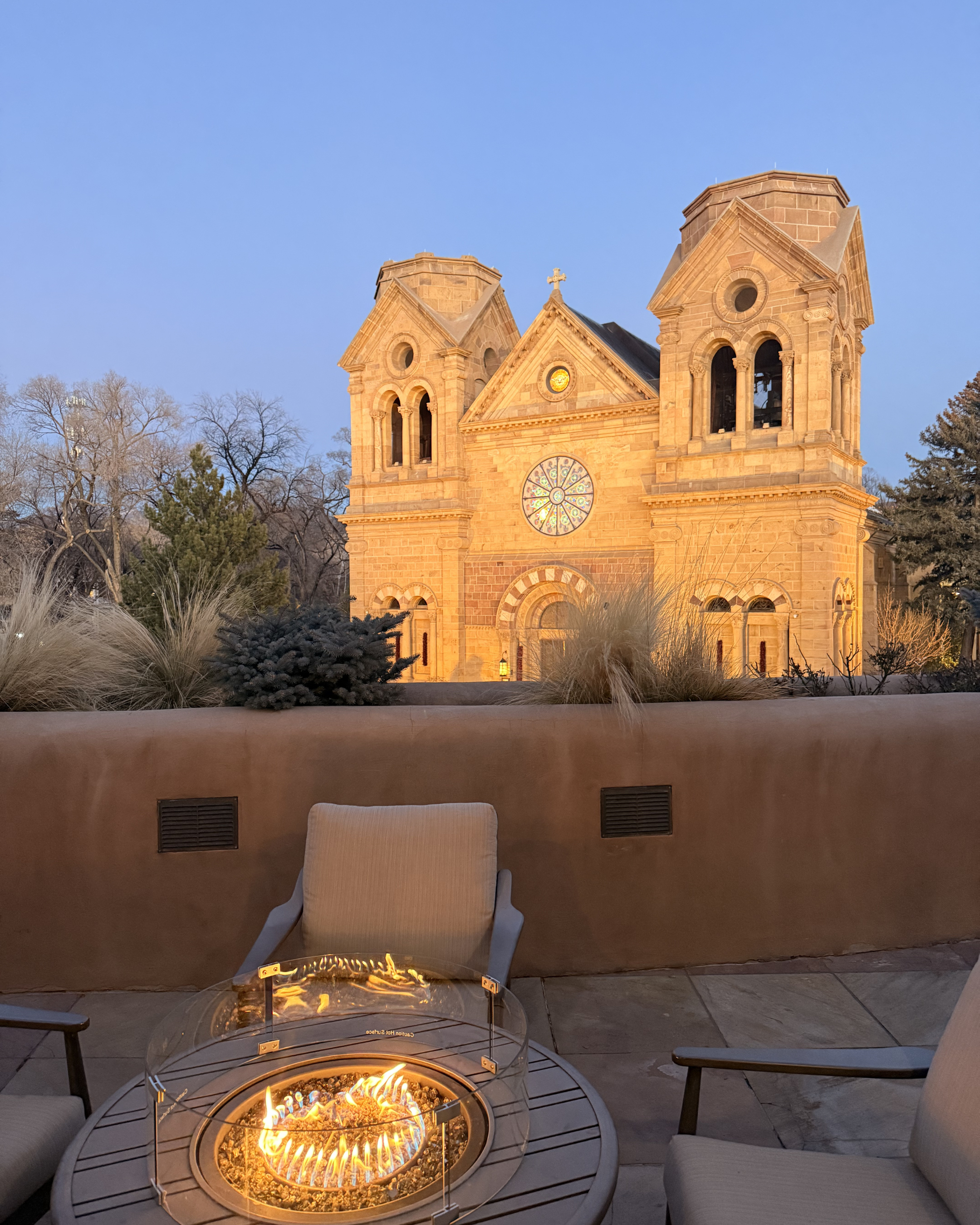 An outdoor seating area with cushioned chairs and tables around a fire pit with flames, set against a backdrop of a church with two towers and a rose window, during dusk or evening in Santa Fe, NM.
