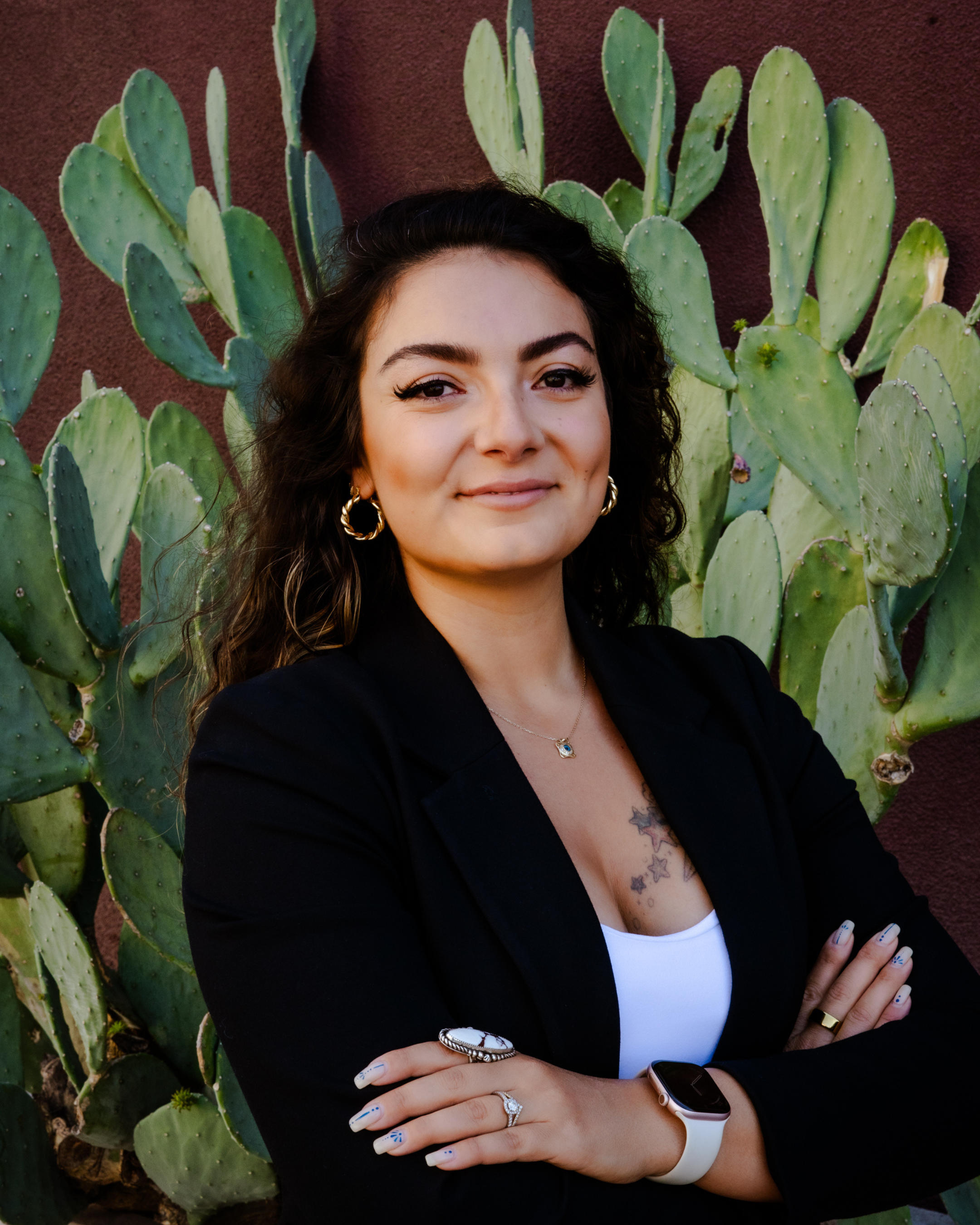 A woman with long curly dark hair and tan skin stands in front of a large cactus with green pads. She is smiling, wearing gold hoop earrings, a black blazer, a white top, a ring, a watch, and has a tattoo on her chest.