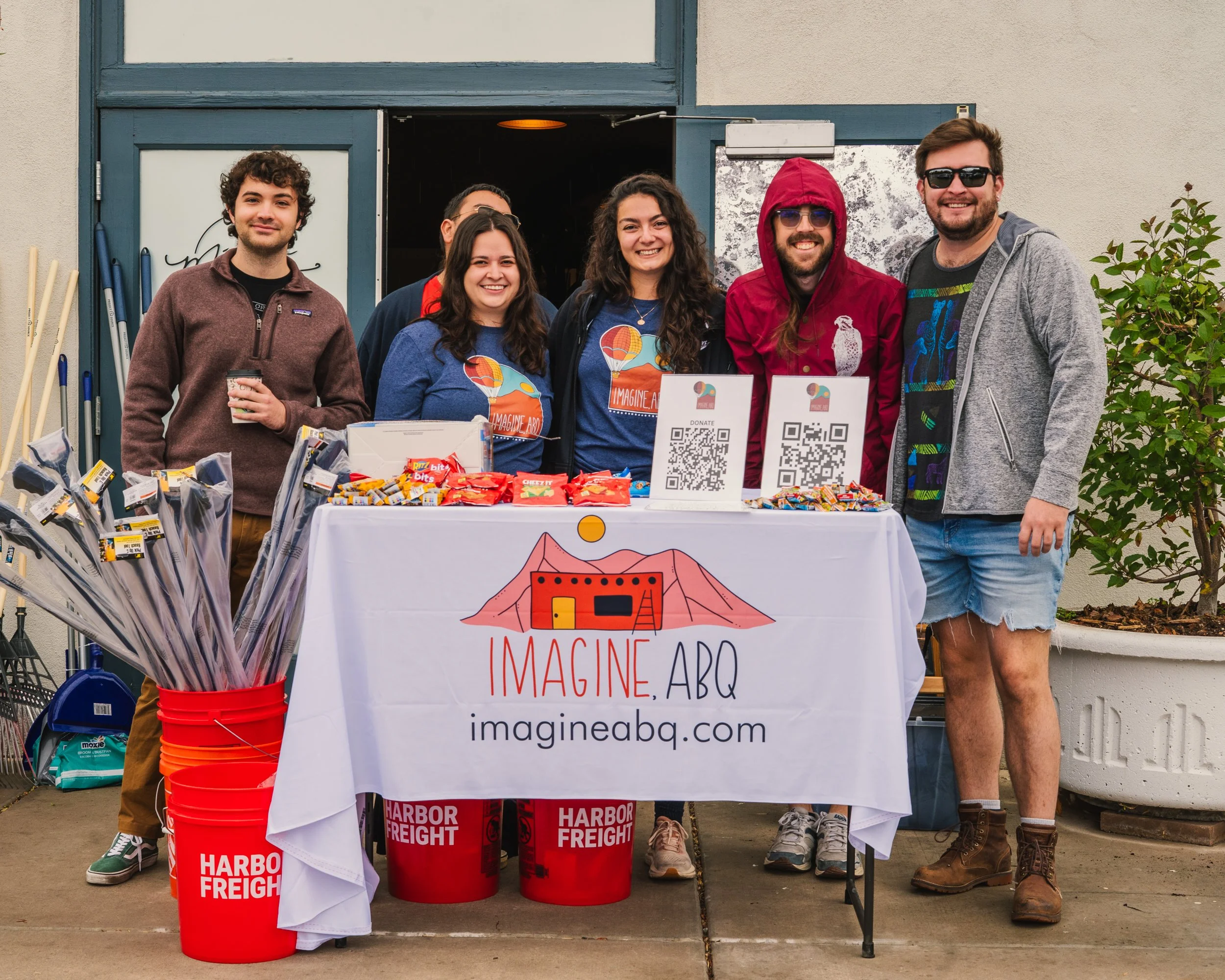 Group of six people standing behind a table with snacks and signs at a charity event, smiling at the camera, outside a building.