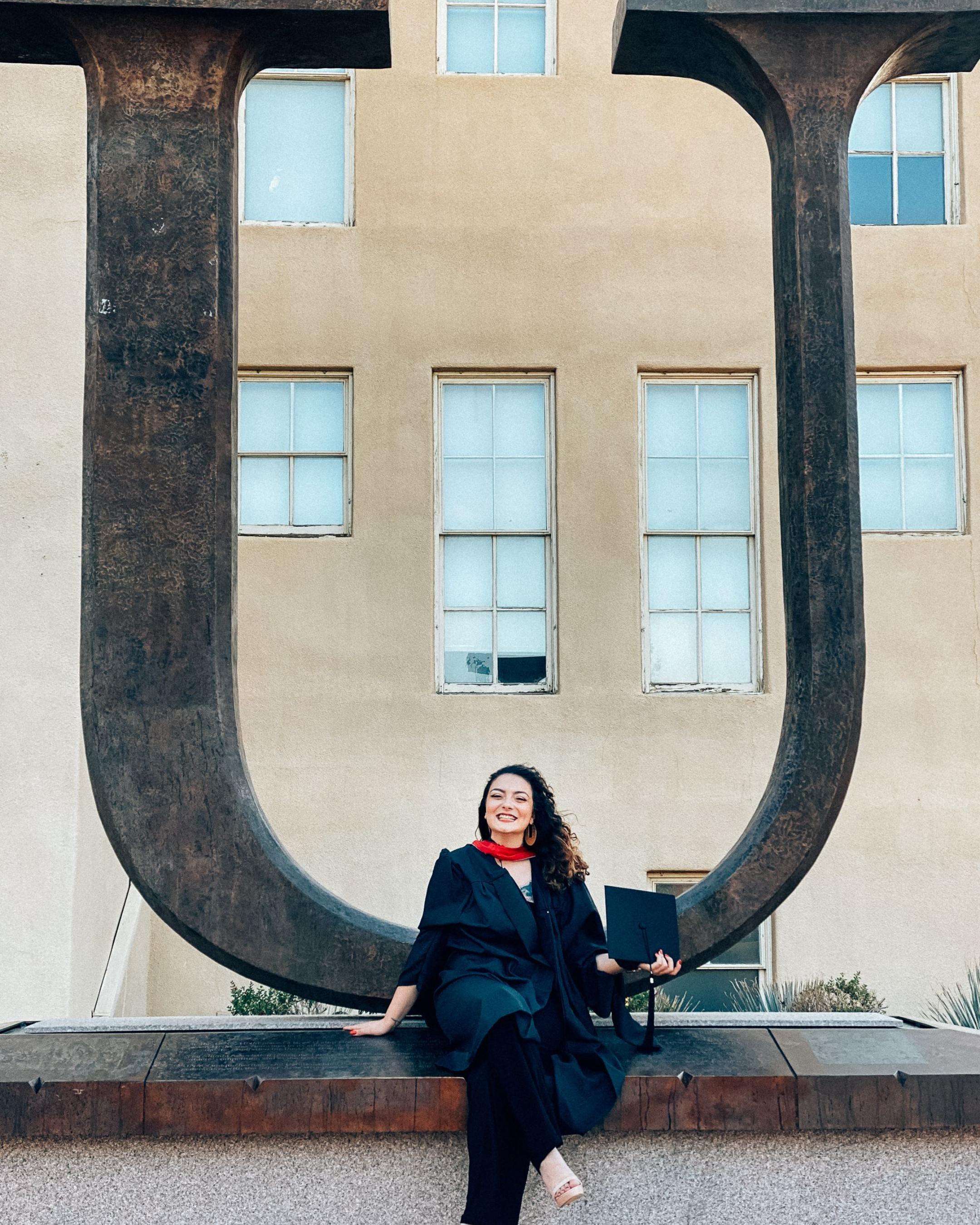 A woman in a graduation gown sitting on a ledge underneath a large metal U-shaped sculpture frame, holding a diploma, with a building with windows in the background.