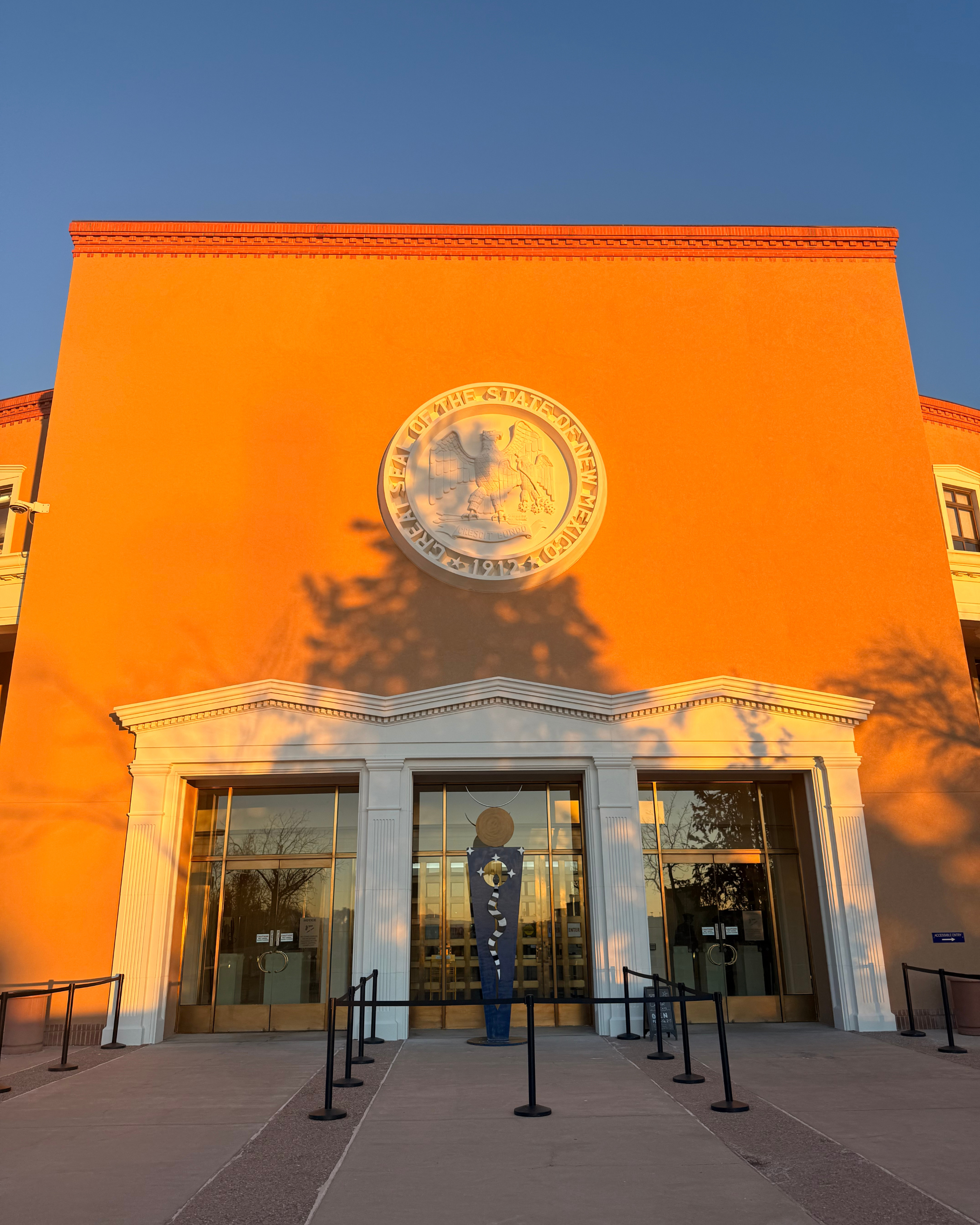 A historic building, the New Mexico State Capitol in Santa Fe, with a white-framed entrance door, large windows, a large emblem of the United States Flag with an eagle, illuminated by warm sunlight.