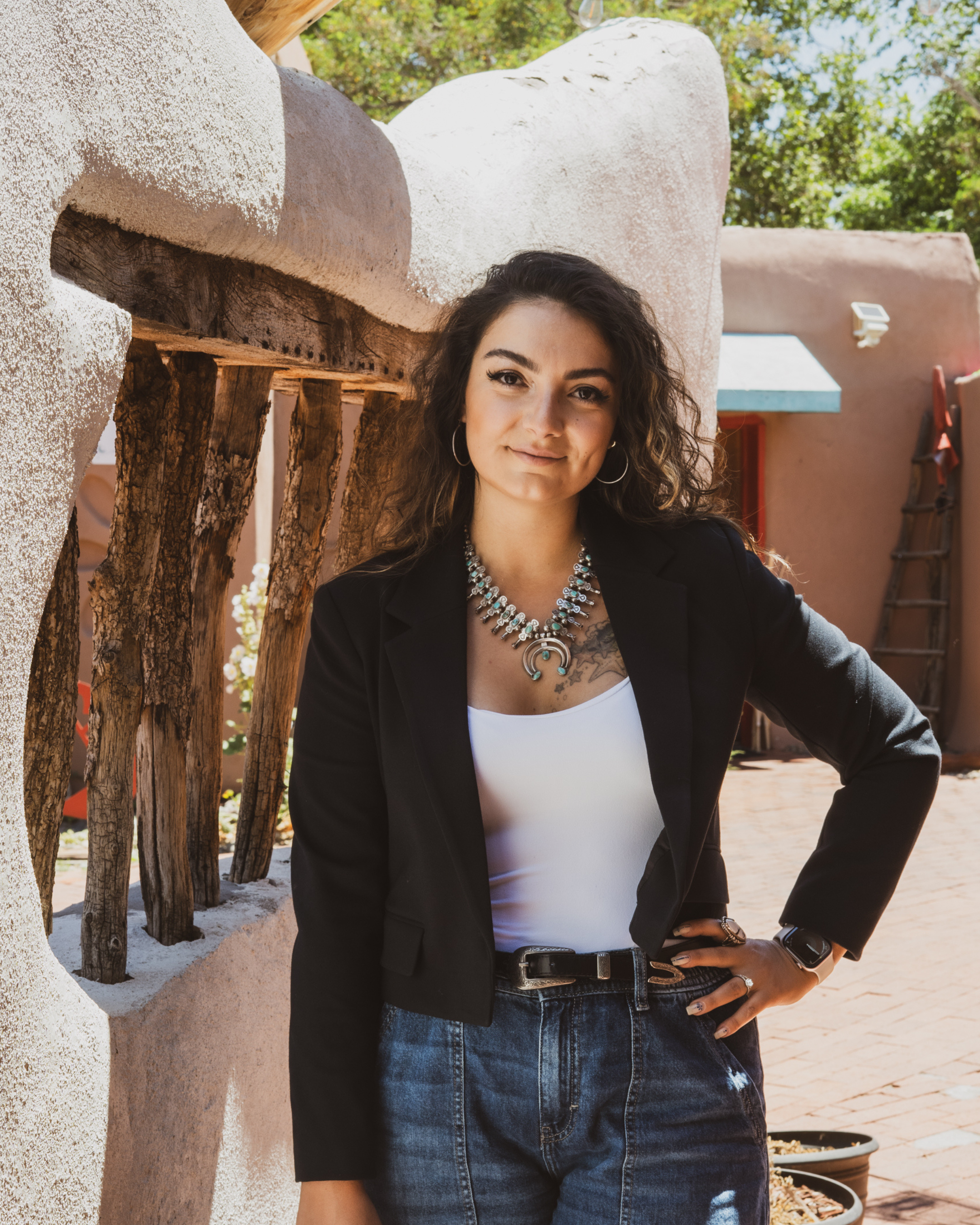 A woman with curly hair wearing a black blazer, white top, squash blossom neckalce, and jeans, standing outdoors near a rustic wooden fence and a stucco building with trees in the background.