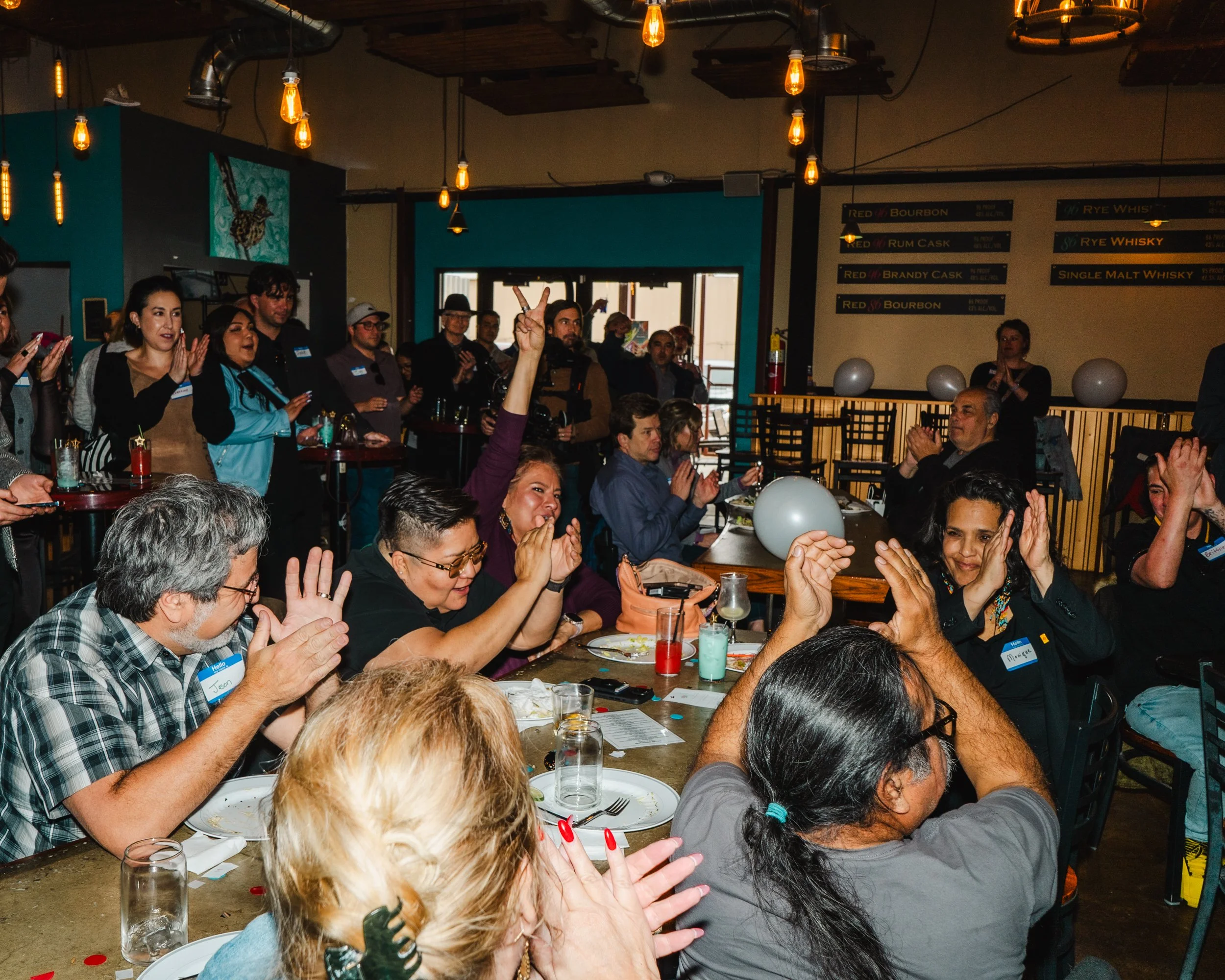 People gathered around tables in a dimly lit restaurant or bar, some clapping and raising hands, with hanging Edison bulbs and a menu board on the wall in the background.