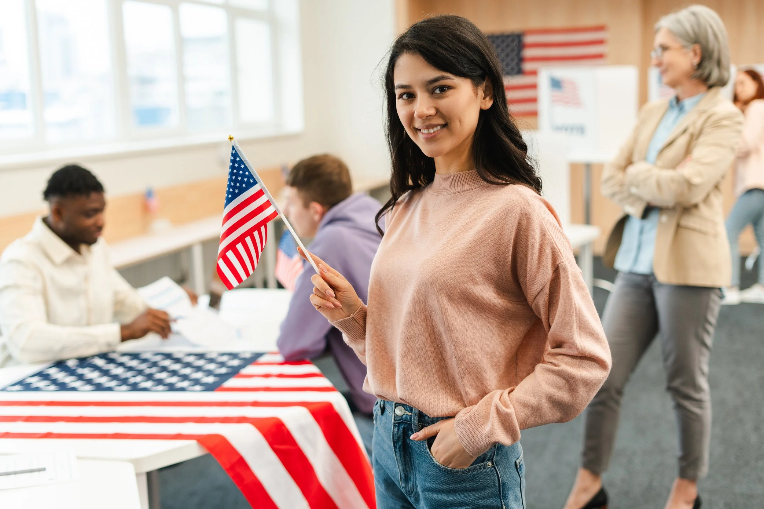 A young woman with dark hair and a pink sweater smiling at the camera while holding a small American flag at an indoor event decorated with American flags and patriotic tablecloths, with other people in the background.