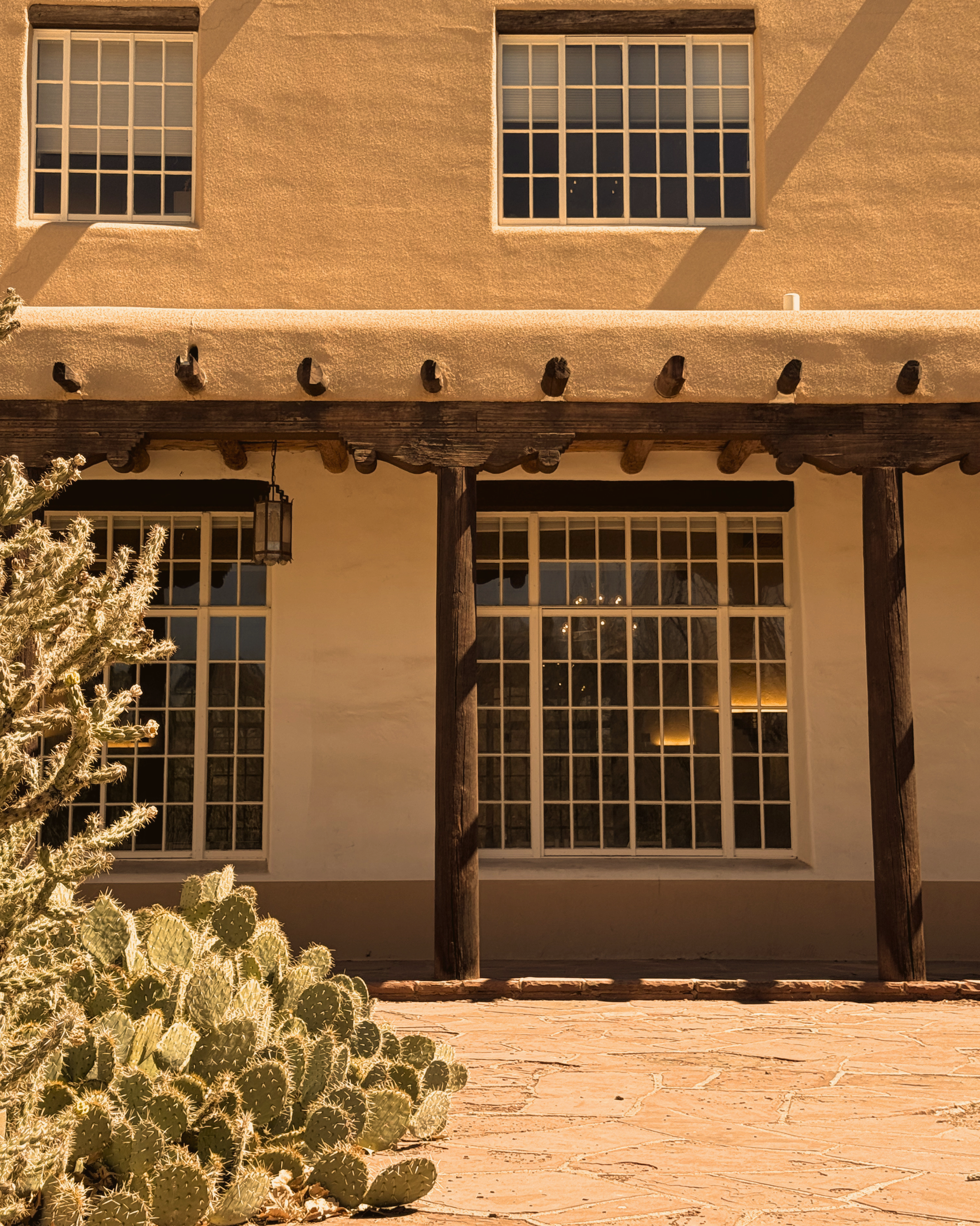 Exterior view of a building with two tall windows with white grid panes, a stucco wall, and a rustic wooden porch with beams. In the foreground, there are cactus plants.