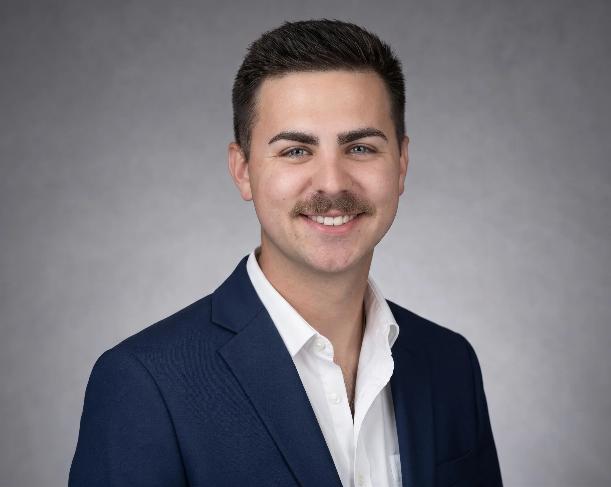 A young man with dark hair, a mustache, and blue eyes, smiling and wearing a navy blue suit jacket over a white shirt, against a gray background.