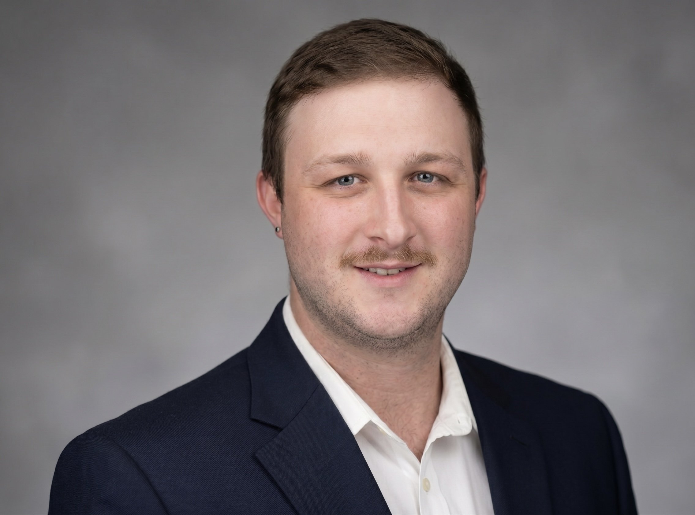 Close-up of a young man with short brown hair, blue eyes, and a slight beard, wearing a dark suit jacket and white dress shirt against a plain gray background.