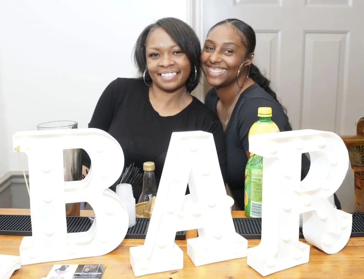 Two women smiling behind illuminated white letters spelling 'BAR' on a bar counter at an indoor social gathering.