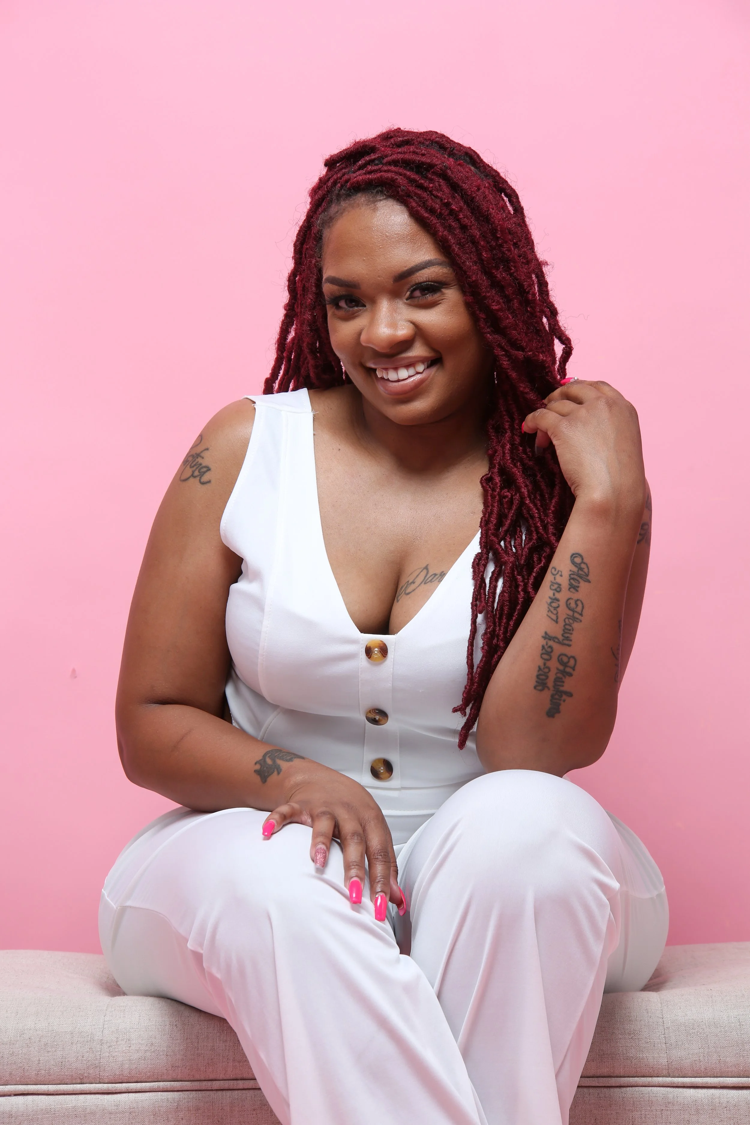 A smiling woman with red dreadlocks sitting against a pink background, wearing a white sleeveless jumpsuit with brown buttons, showing tattoos on her arm and chest, and pink nail polish.