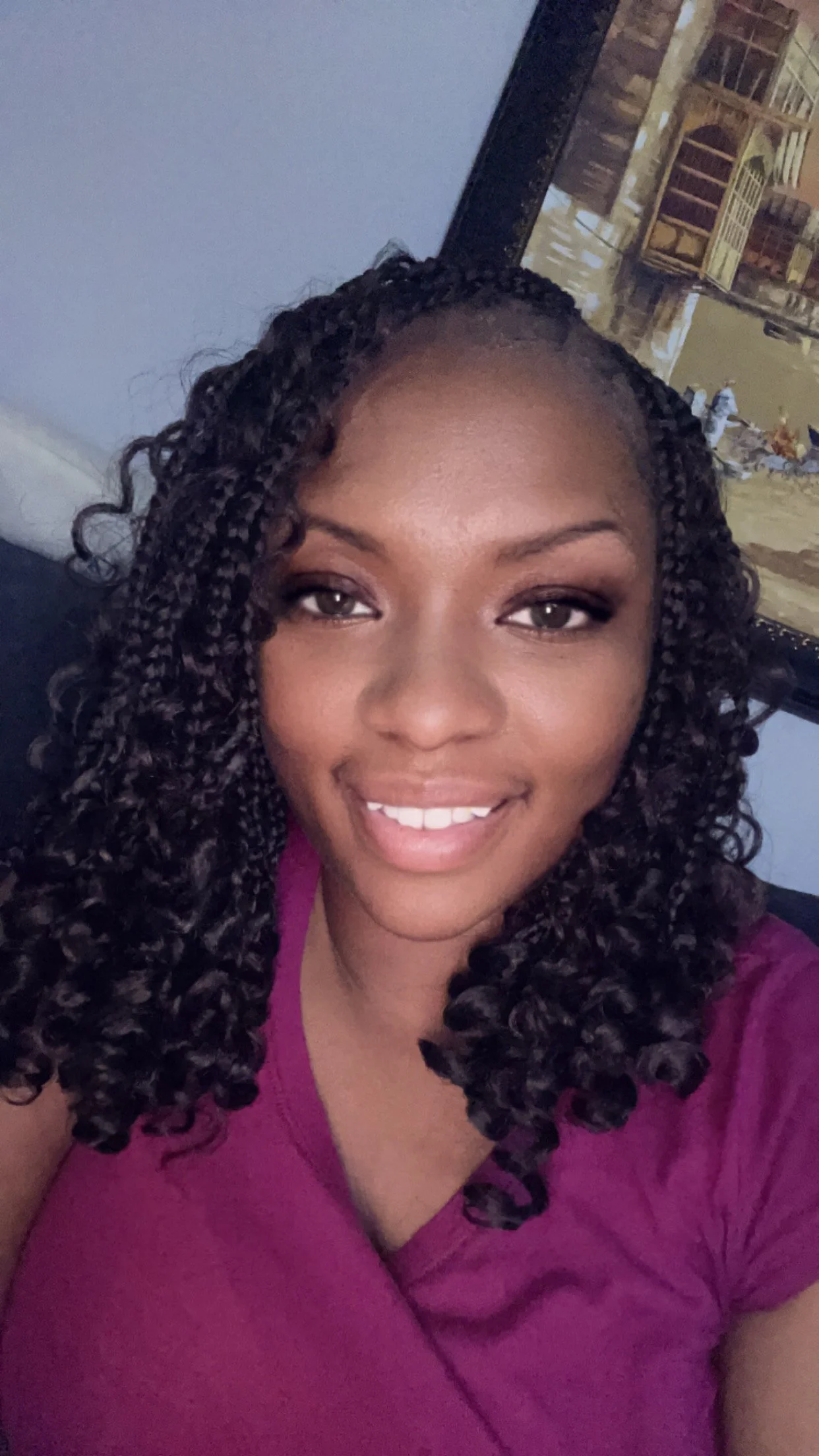 A young woman with curly hair and a bright smile, wearing a purple top, sitting indoors with a framed picture of a cityscape in the background.