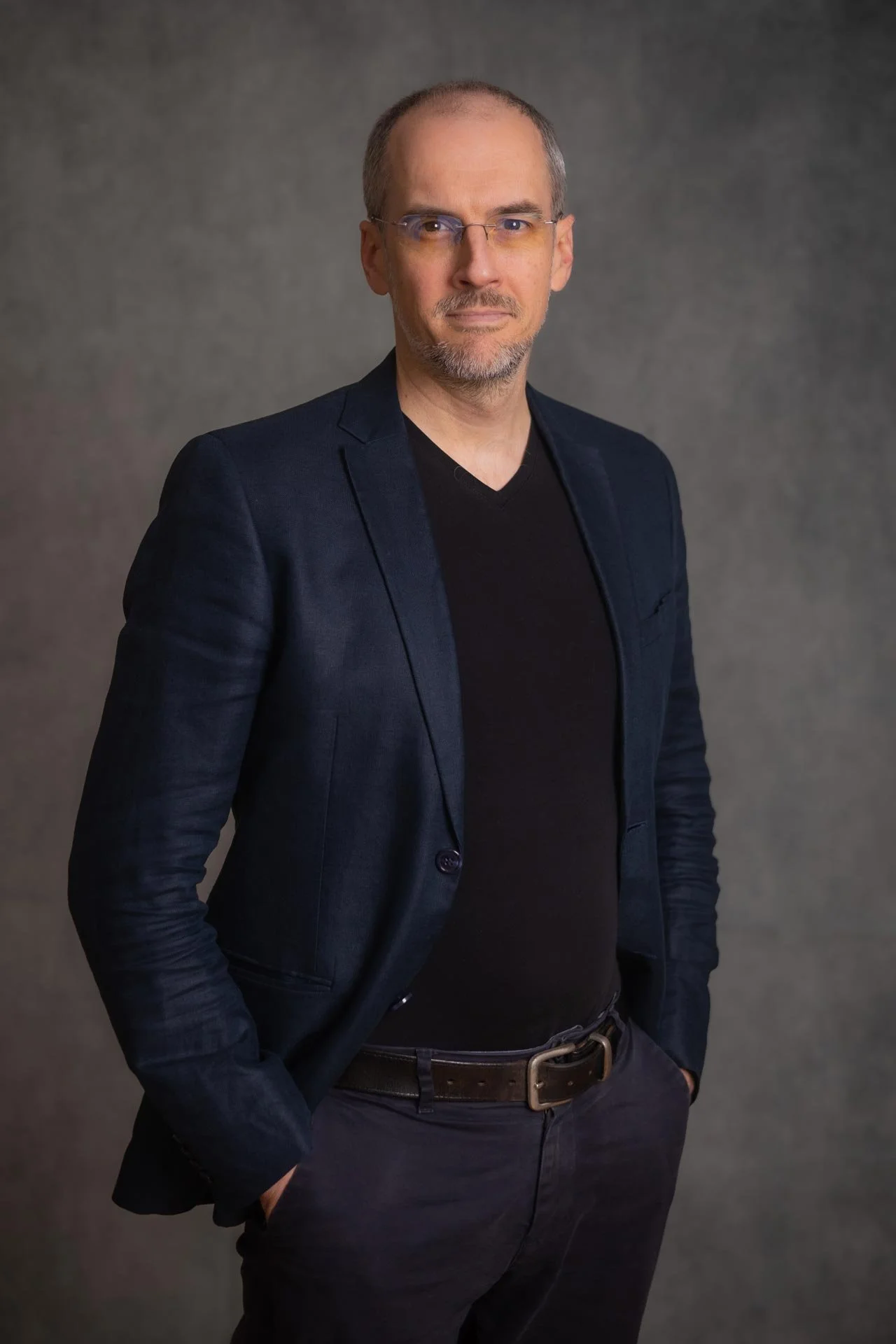 A middle-aged man with glasses and receding hair, wearing a black shirt, navy blazer, and dark pants, standing against a plain gray background with hands in pockets.