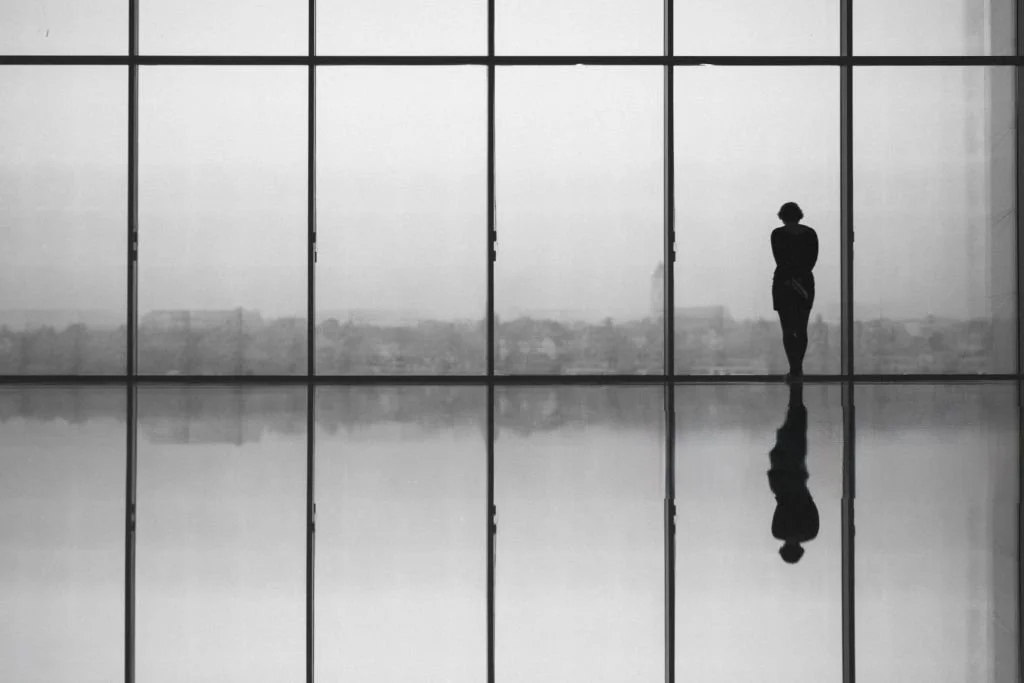 A woman standing alone inside a modern building with large glass windows, overlooking a city skyline in the background. Her reflection is visible on the polished floor.