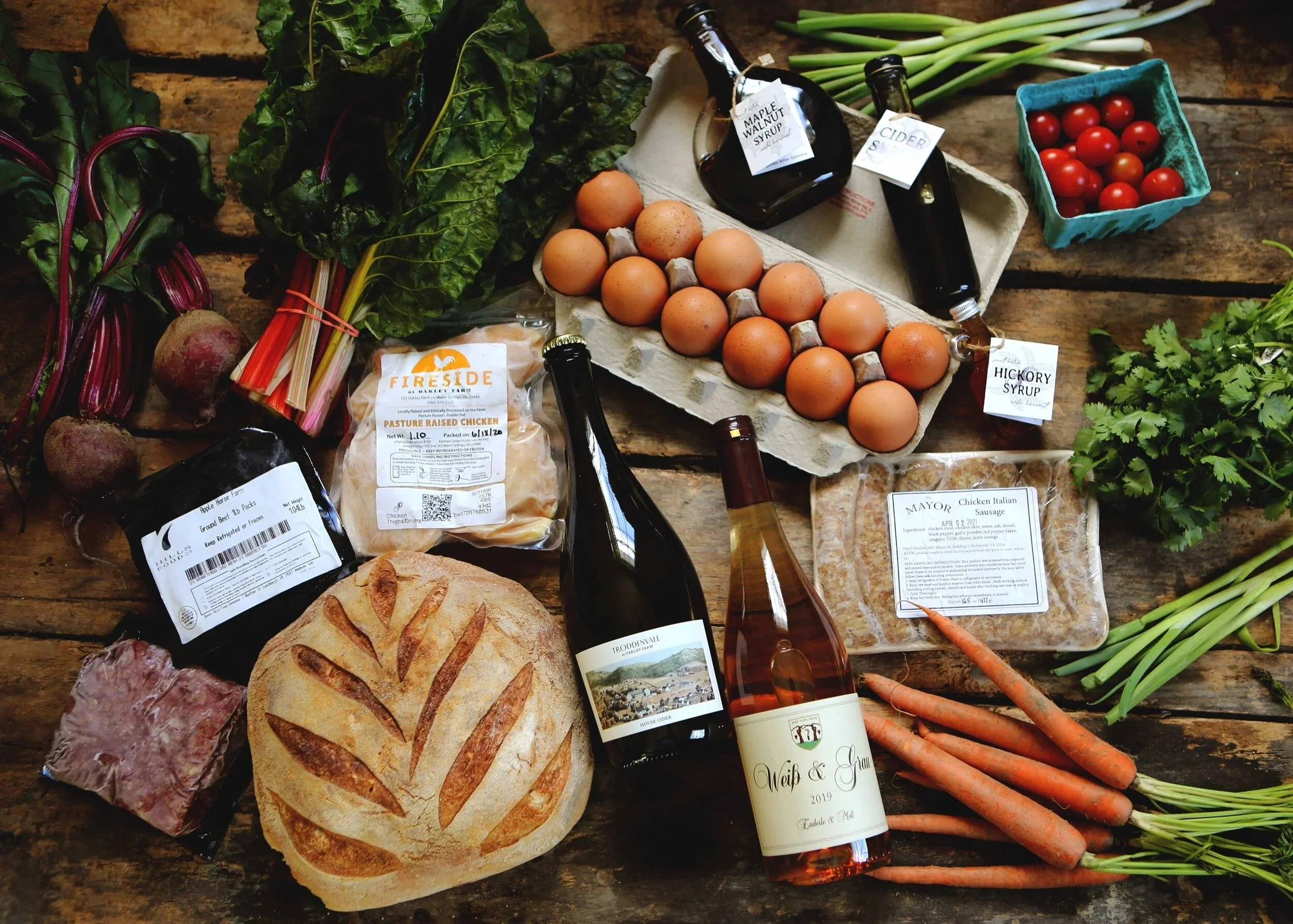 A rustic wooden table displaying fresh vegetables, eggs, bottles of syrup and cider, and a loaf of bread, arranged for a farm or market setting.