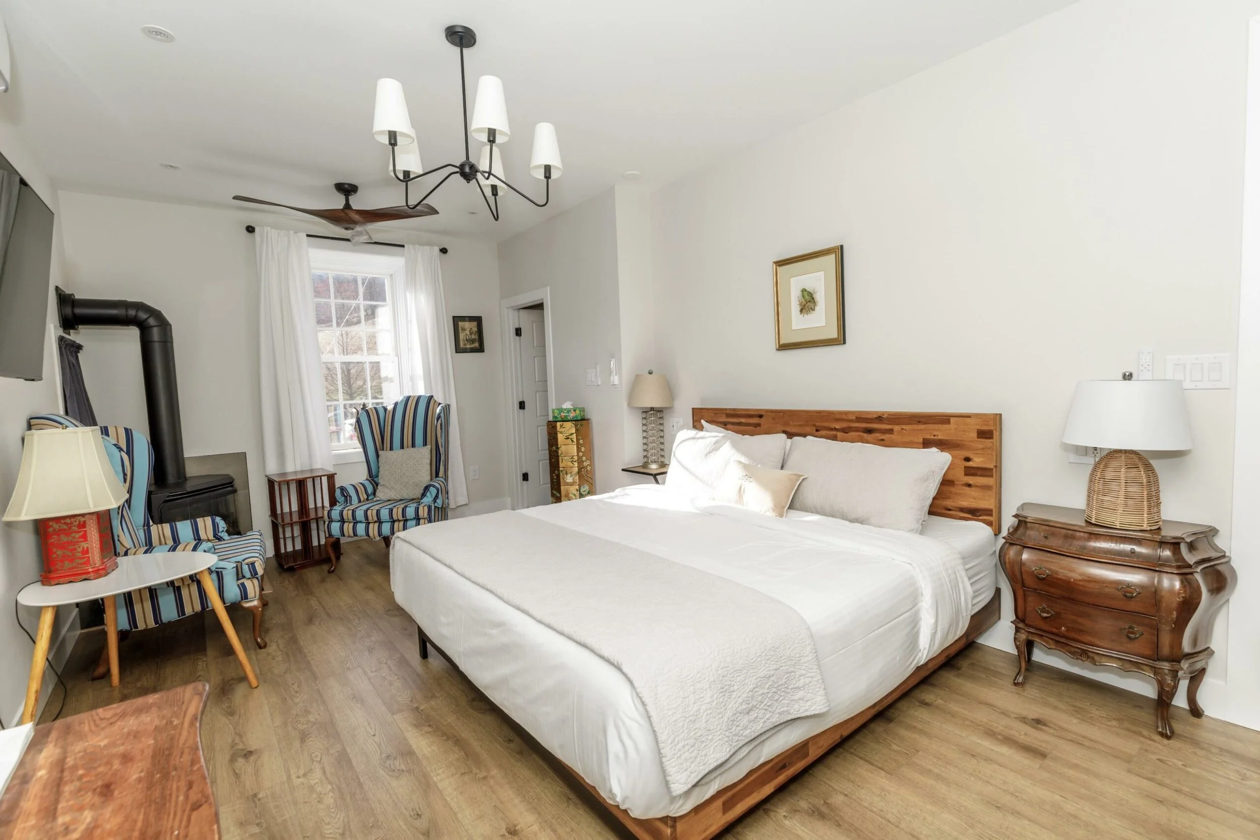 A bedroom with a white bed, wooden headboard, and multiple lamps on nightstands. There are framed artworks on the wall, striped chairs near a window with white curtains, and a wood-burning stove in a corner. The room has wooden flooring and white walls.