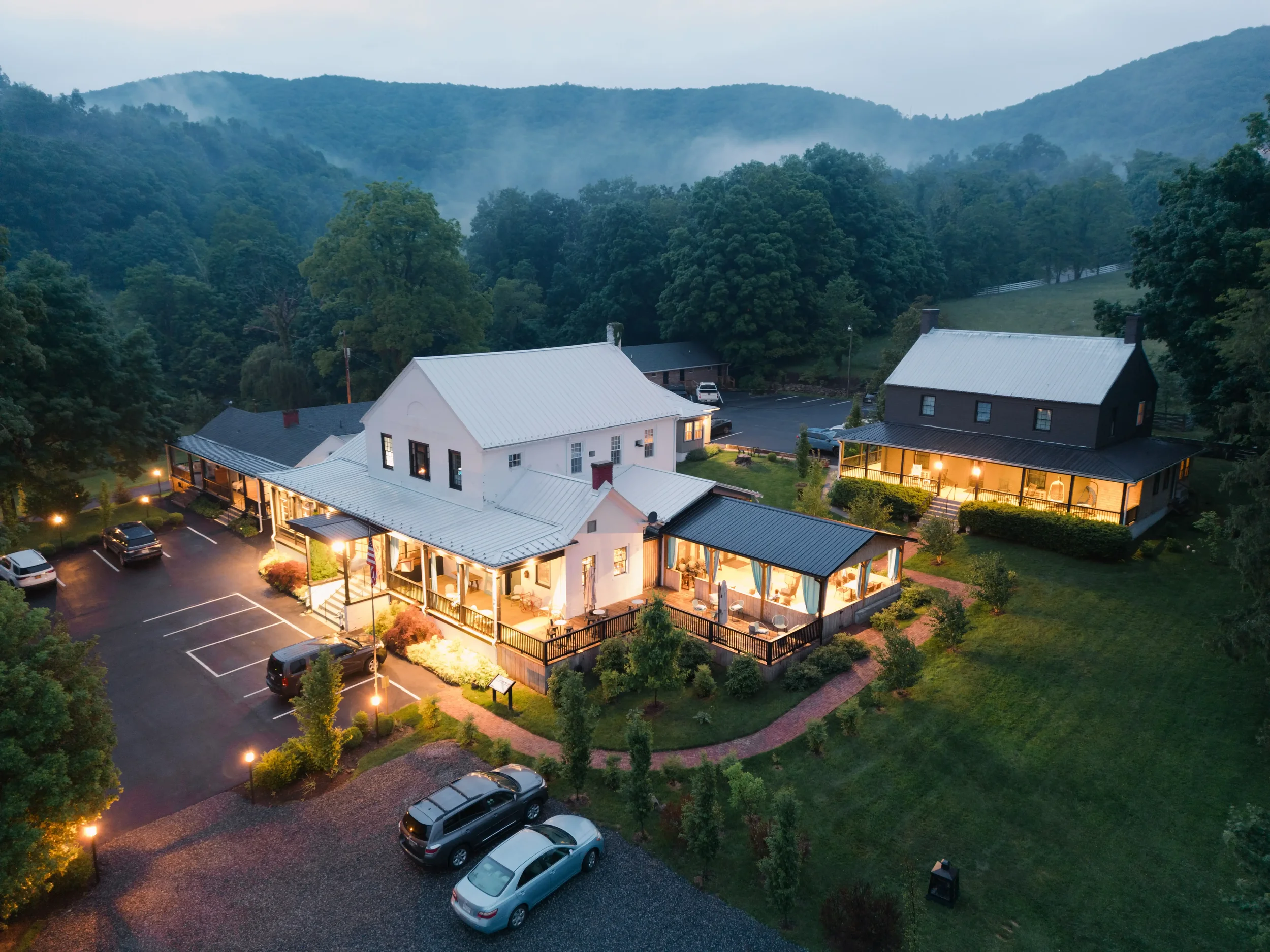 Aerial view of a mountain town at dusk with houses, parked cars, and trees in the foreground, misty mountains in the background.