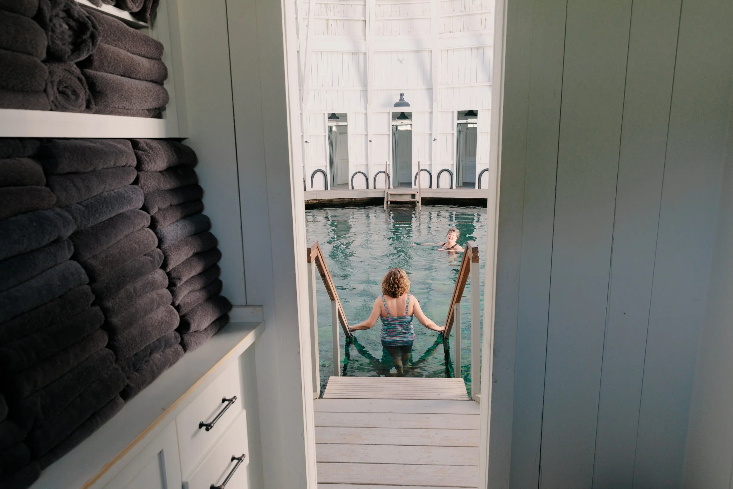 View through a closet door showing two women in swimsuits exiting into a dock with water, stairs, and white walls with mirrors, with one woman in water and one on stairs.