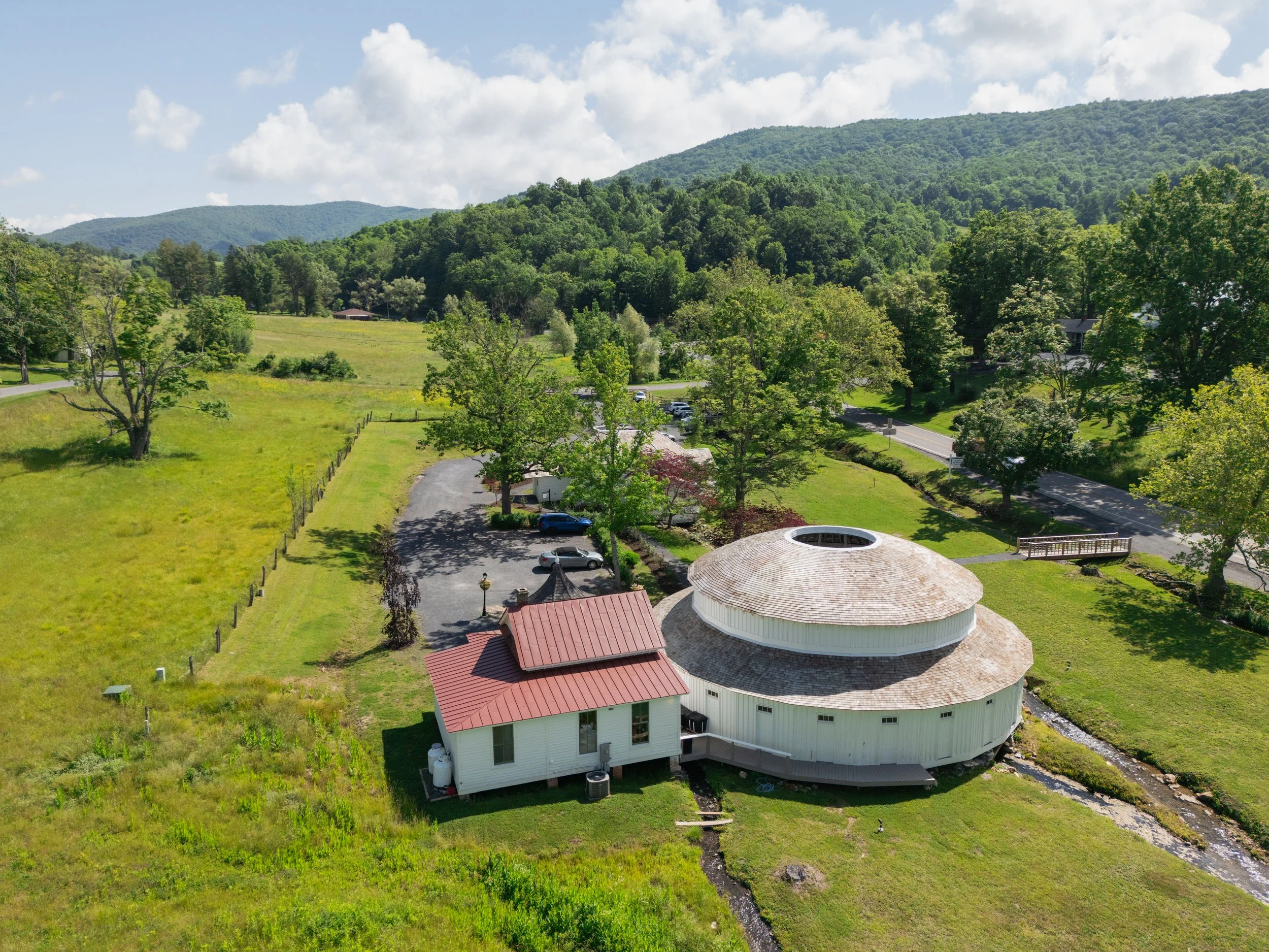 A white circular barn with a conical roof and red accents, situated in a green rural landscape with trees, a road, and mountain hills in the background.