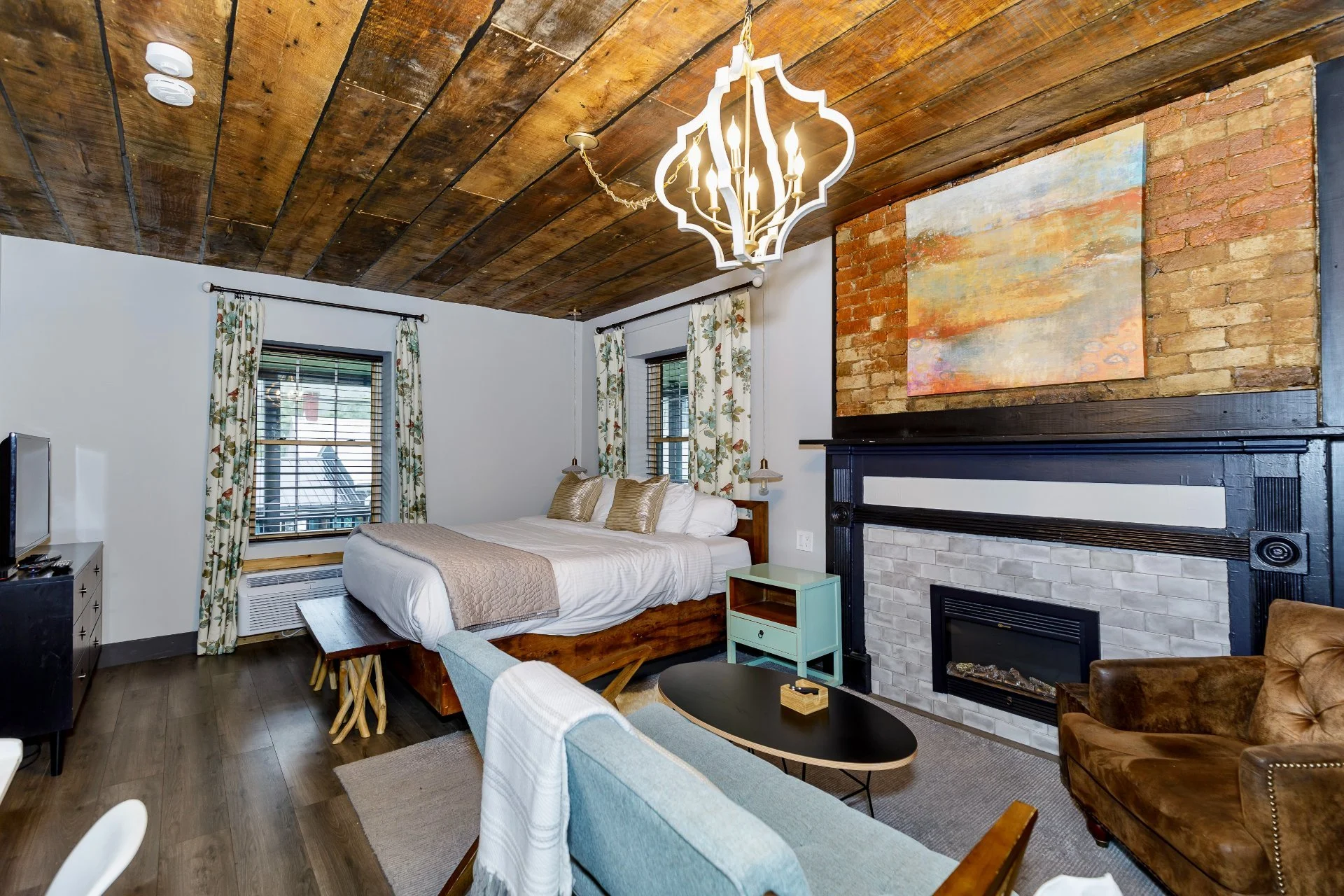 A cozy bedroom with a wooden ceiling, a wall with exposed brick, and a fireplace. The bed has white linens and gold pillows, and there are windows with floral curtains. A chandelier hangs from the ceiling, and there's a small mint-colored side table next to the bed. A leather armchair and a round black coffee table are also visible.