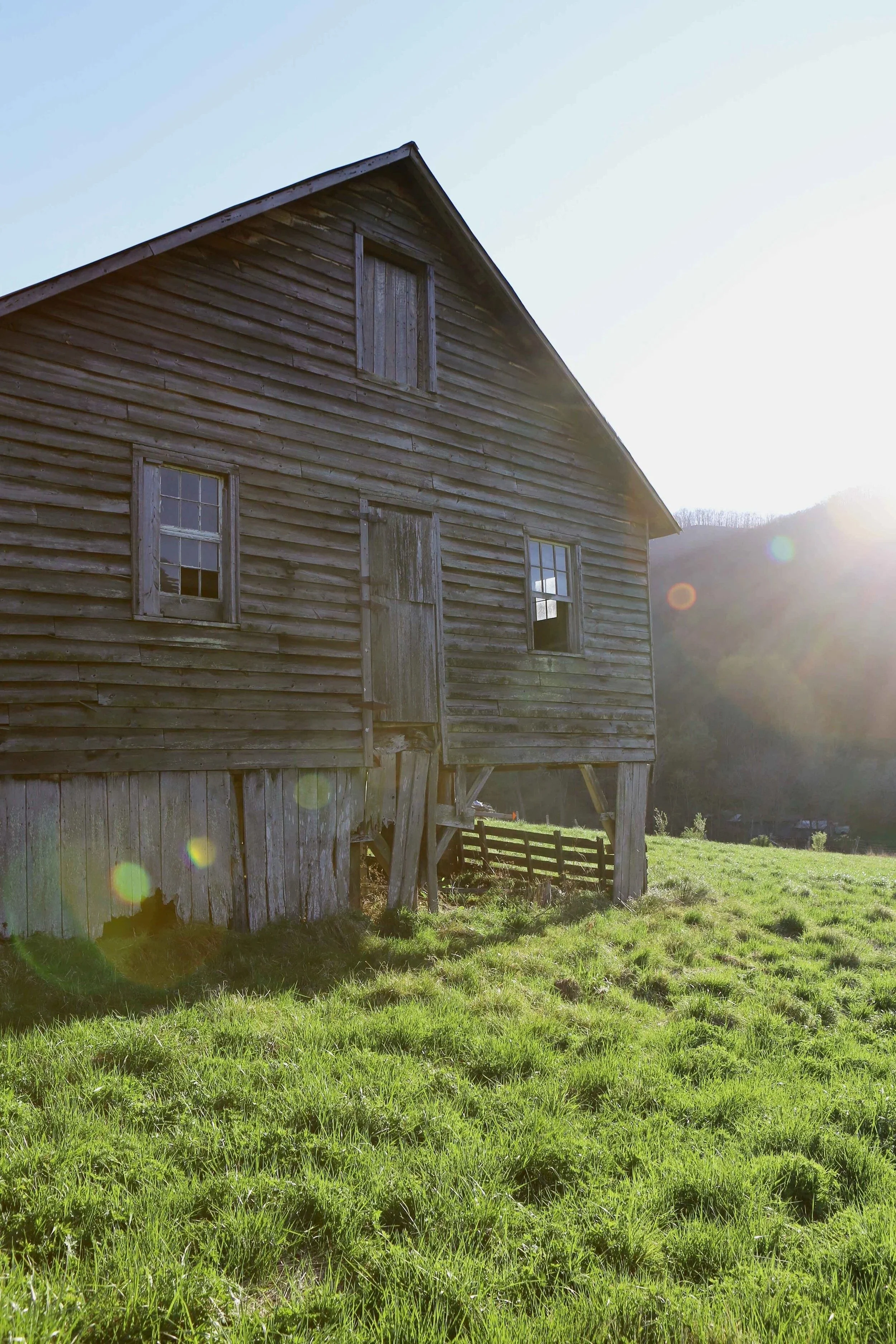 An old wooden barn with boarded-up windows and a weathered exterior, situated in a lush green field with sunlight shining from the right side of the image.